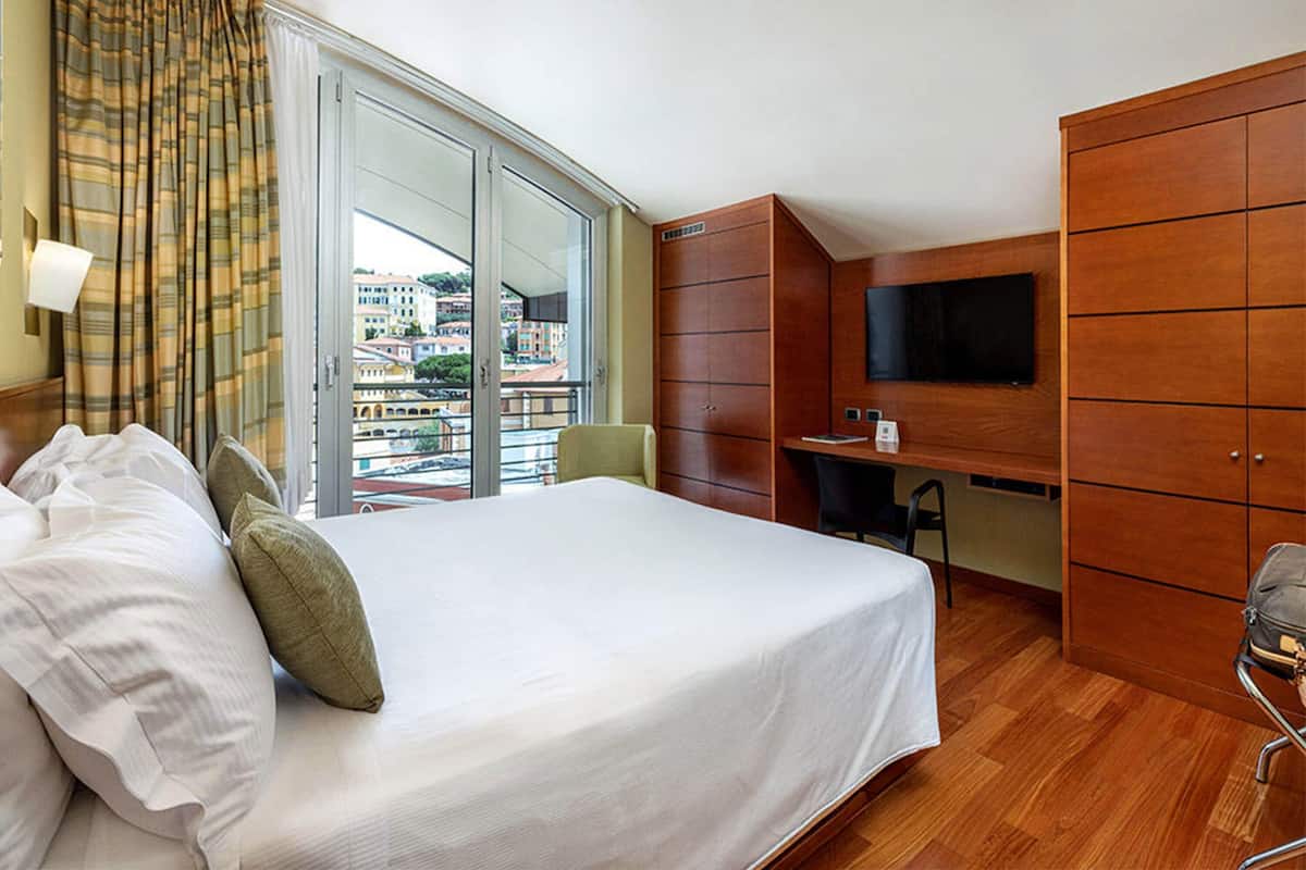 A hotel room with a bed, a desk, a chair, a TV, and a view of buildings through a large window.