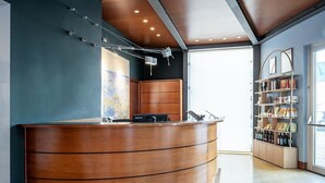 A reception desk with a curved design, a wooden cabinet, and a bookshelf with various items. - B&B HOTEL Imperia Rossini al Teatro (Imperia)