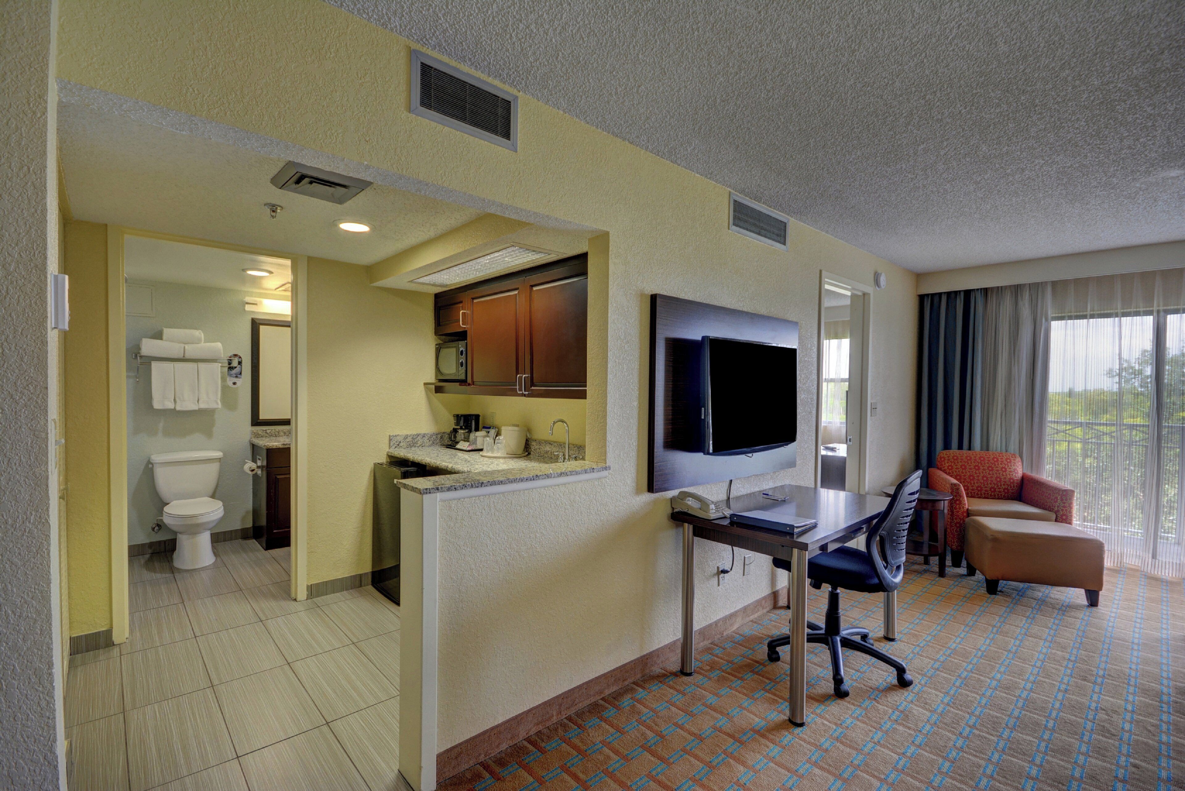 Habitación de hotel con kitchenette, televisor, escritorio, silla y ventana con cortinas.