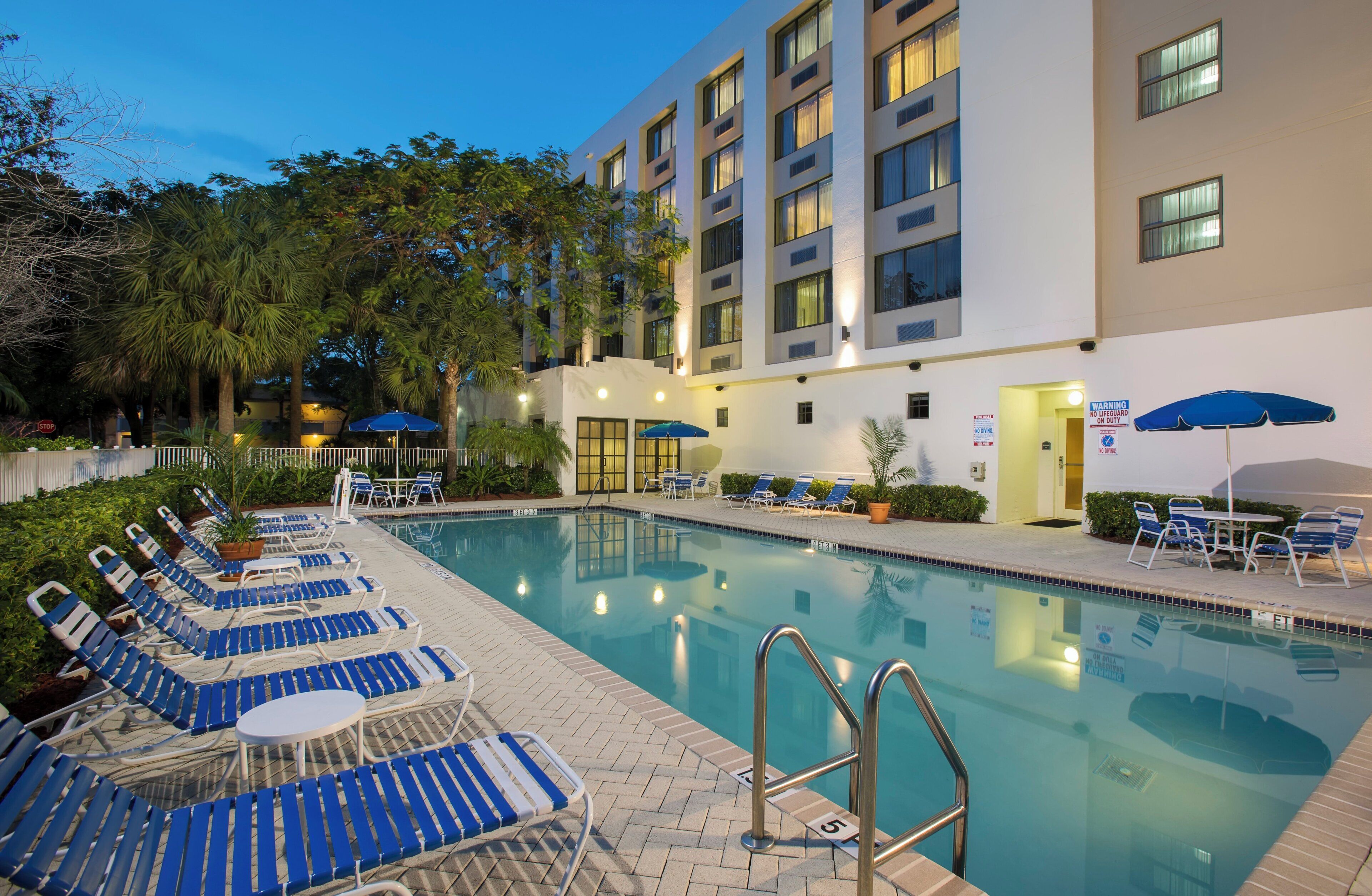 Zona de piscina con sillones de descanso y sombrillas, rodeada por un edificio de varios pisos y ventanas.