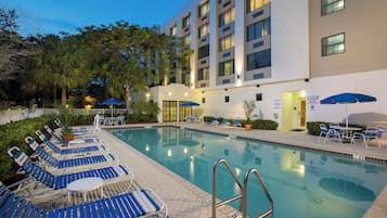 Zona de piscina con sillones de descanso y sombrillas, rodeada por un edificio de varios pisos y ventanas.
