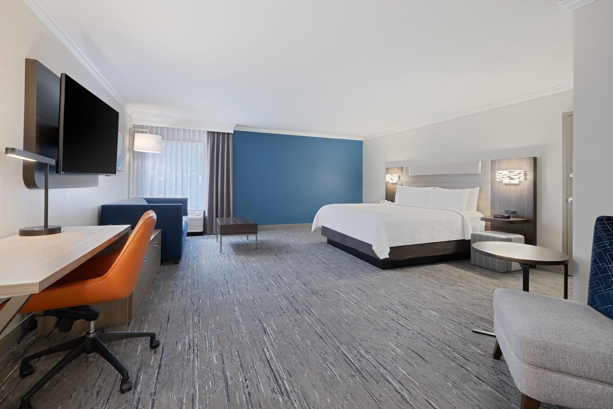 A hotel room with a bed, a desk, an orange chair, a TV, and a small table.