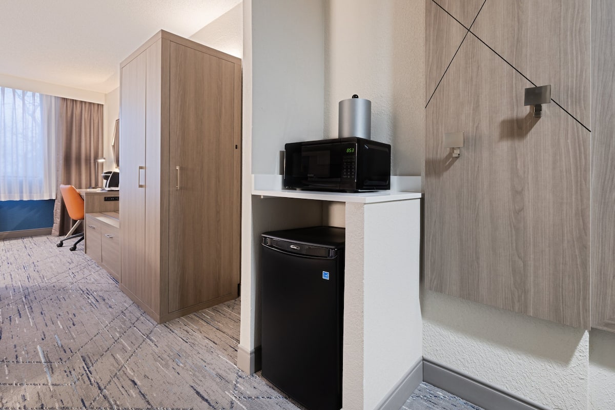A compact hotel room with a kitchenette, a wooden wardrobe, a small desk with a chair, and a view through a window with curtains.