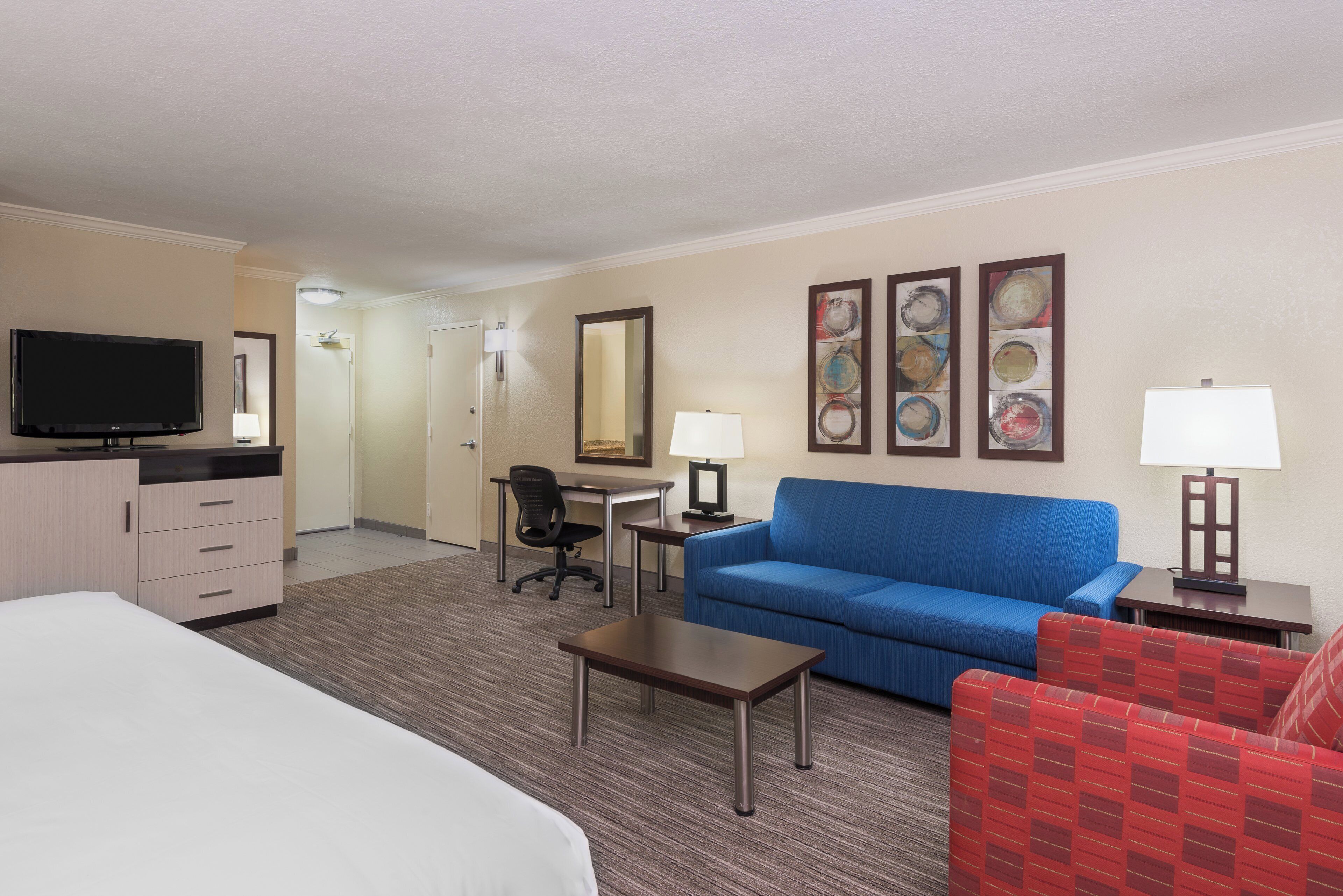 Quarto de hotel com cama, escrivaninha, sofá azul, poltrona vermelha, televisão e obras de arte nas paredes.