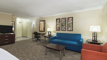 A hotel room with a bed, a blue sofa, a red armchair, a desk with a chair, a television, and artwork on the wall.