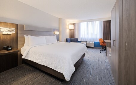A modern hotel room with a large bed, a desk, a chair, and a wardrobe.