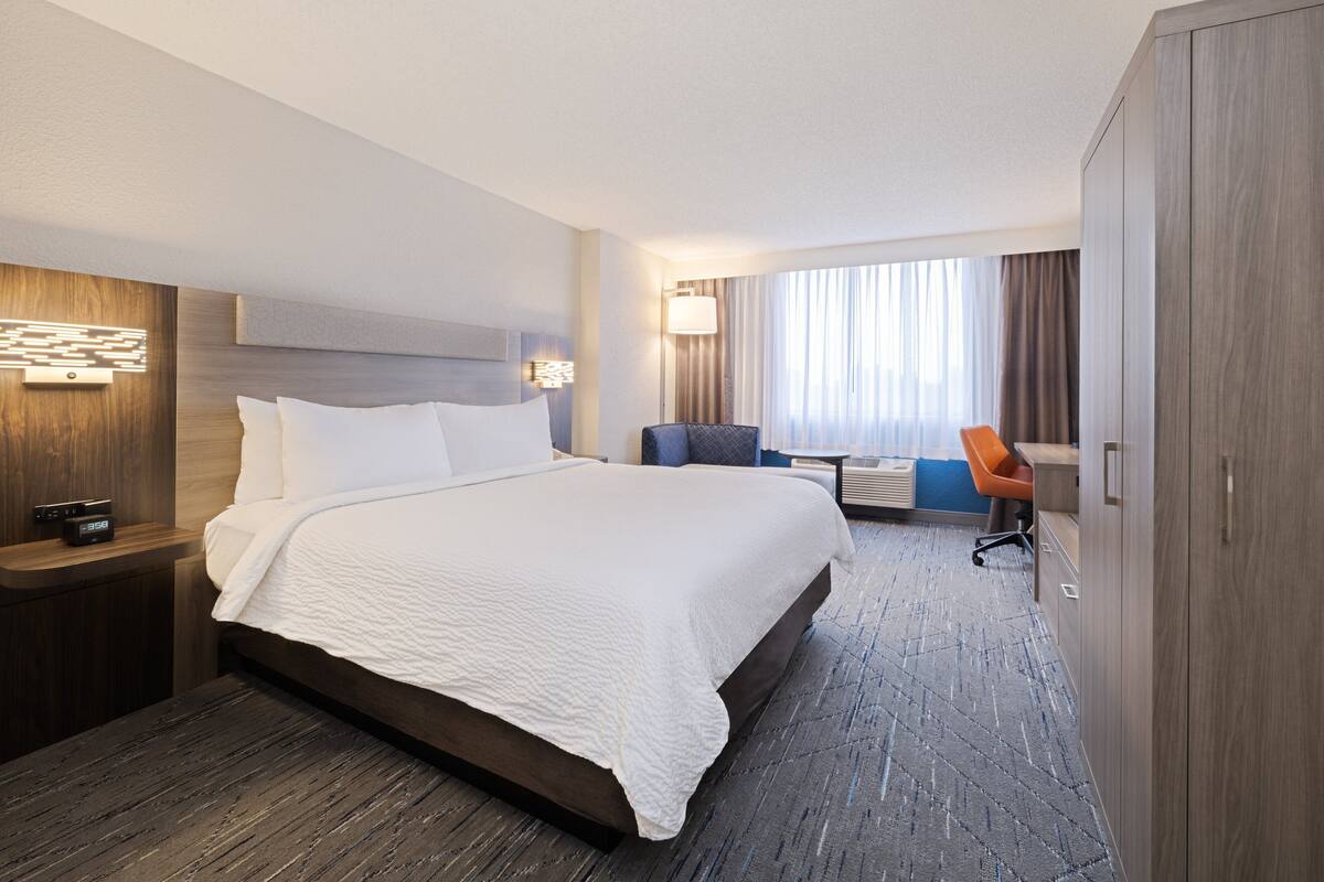 A modern hotel room with a large bed, a desk, a chair, and a wardrobe.