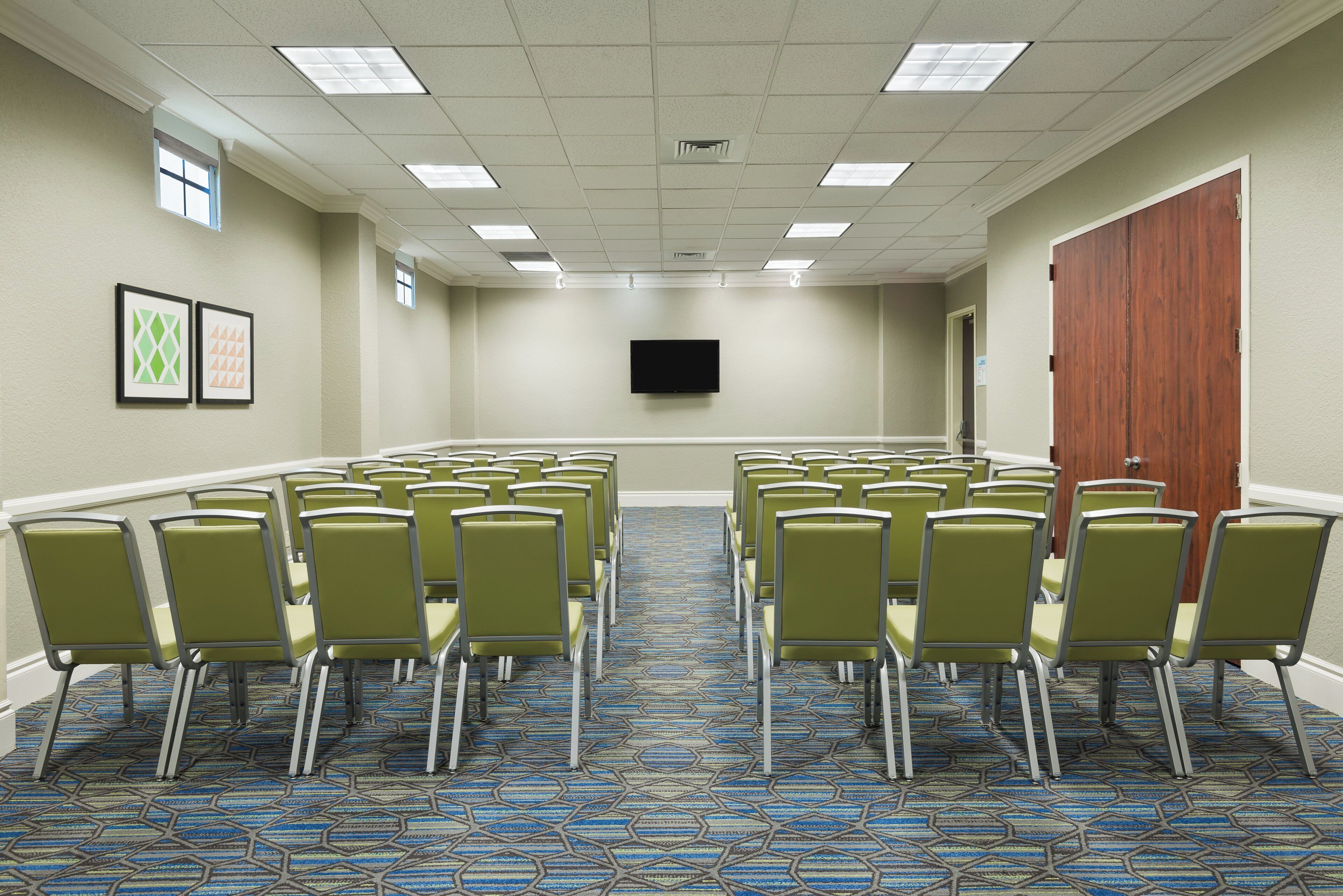 A conference room with rows of chairs, a flat-screen TV on the wall, and a door on the right.
