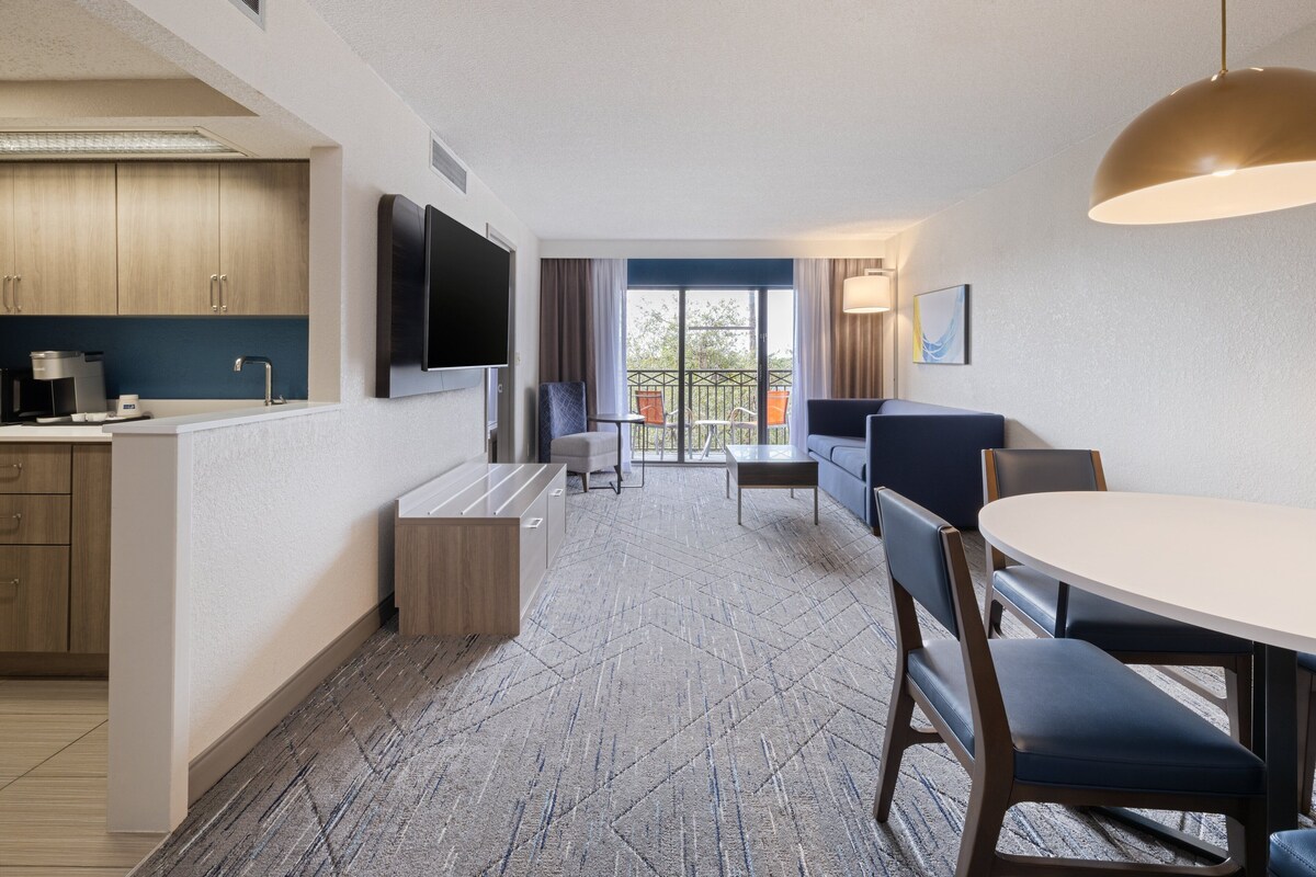 A modern hotel room with a kitchenette, a flat-screen TV, a dining area with a round table and chairs, and a balcony with a view.