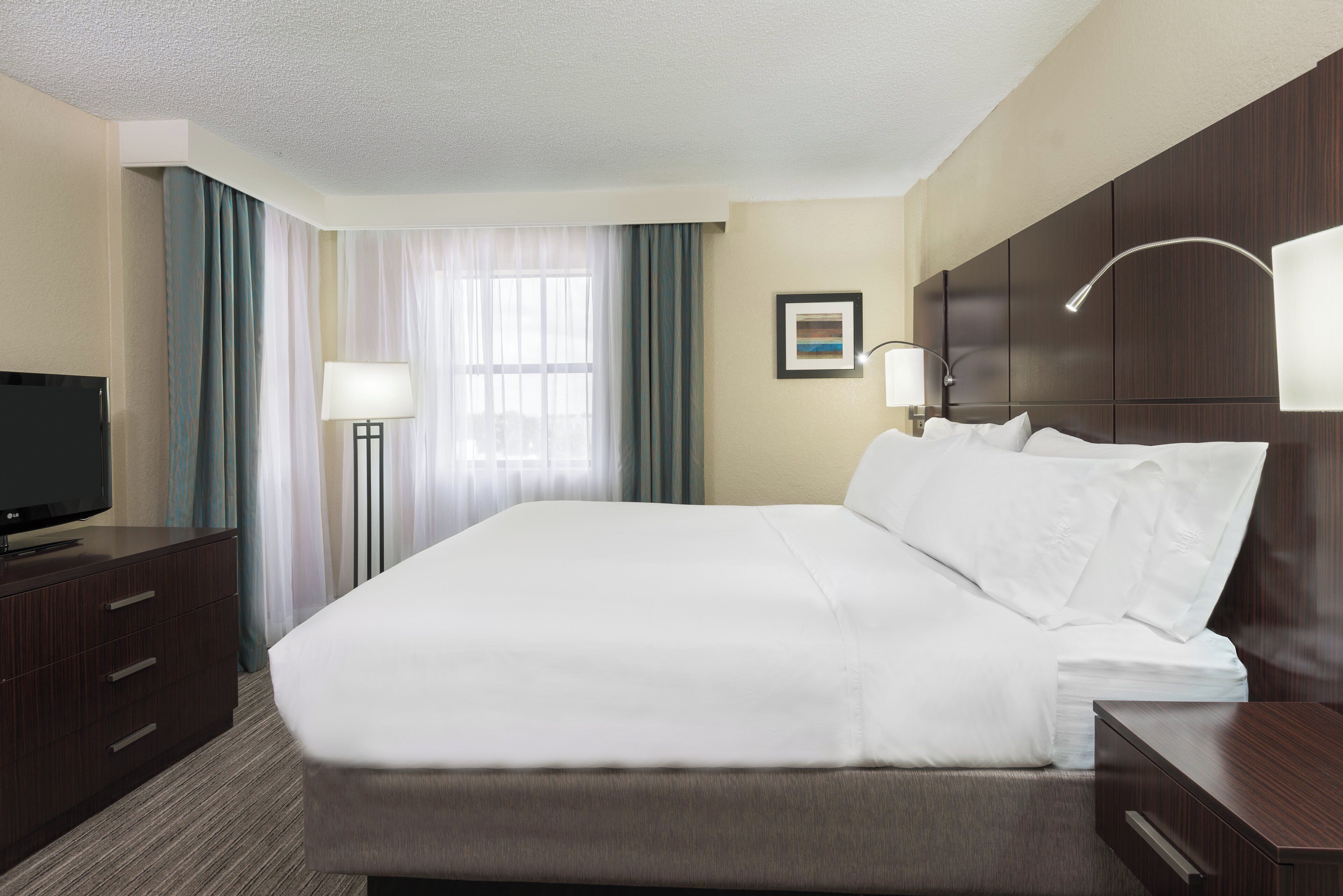A hotel room with a large bed, a flat-screen TV, a desk with a lamp, and a window with curtains.