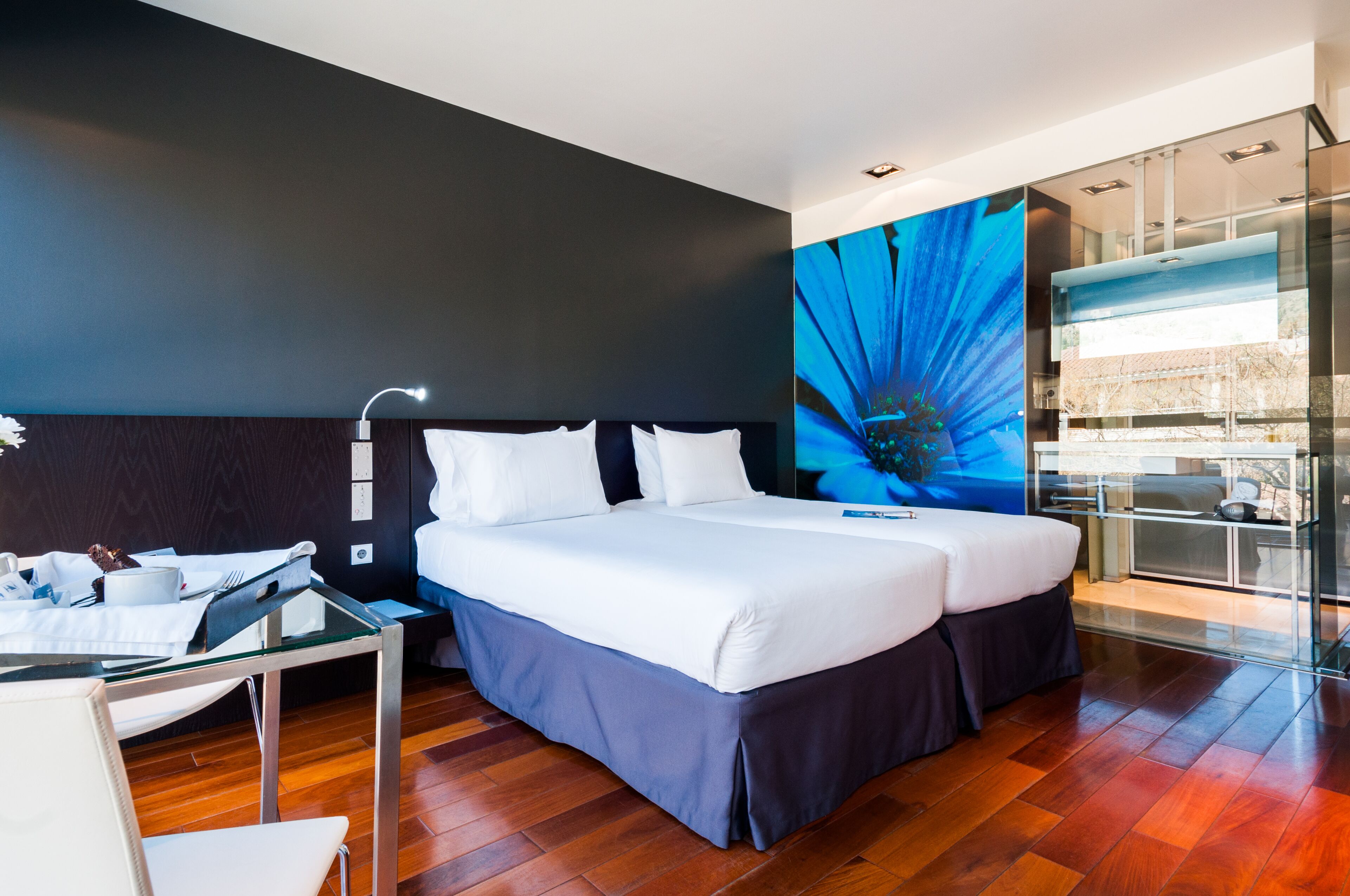 Een moderne hotelkamer met een groot bed, een glazen tafel en uitzicht op de stad door een groot raam.