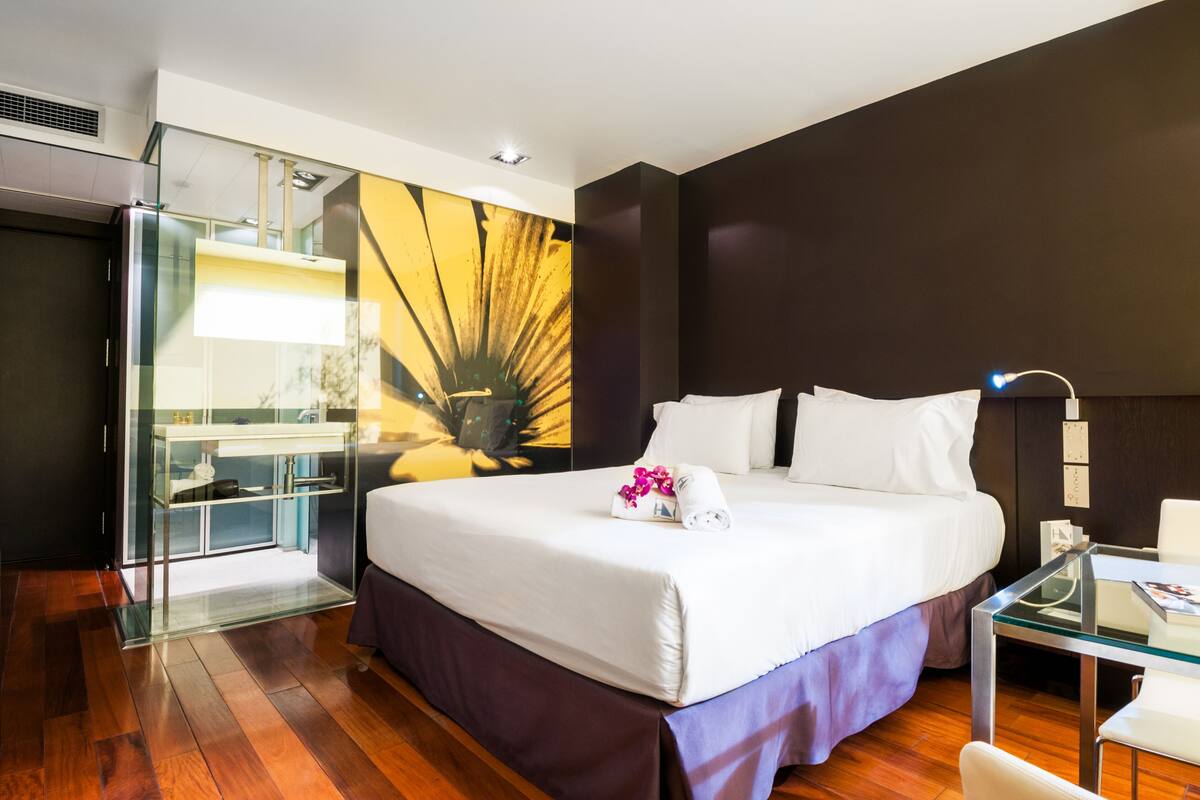 Habitación de hotel moderna con una cama grande, un separador de vidrio, un escritorio y vistas a un paisaje urbano.