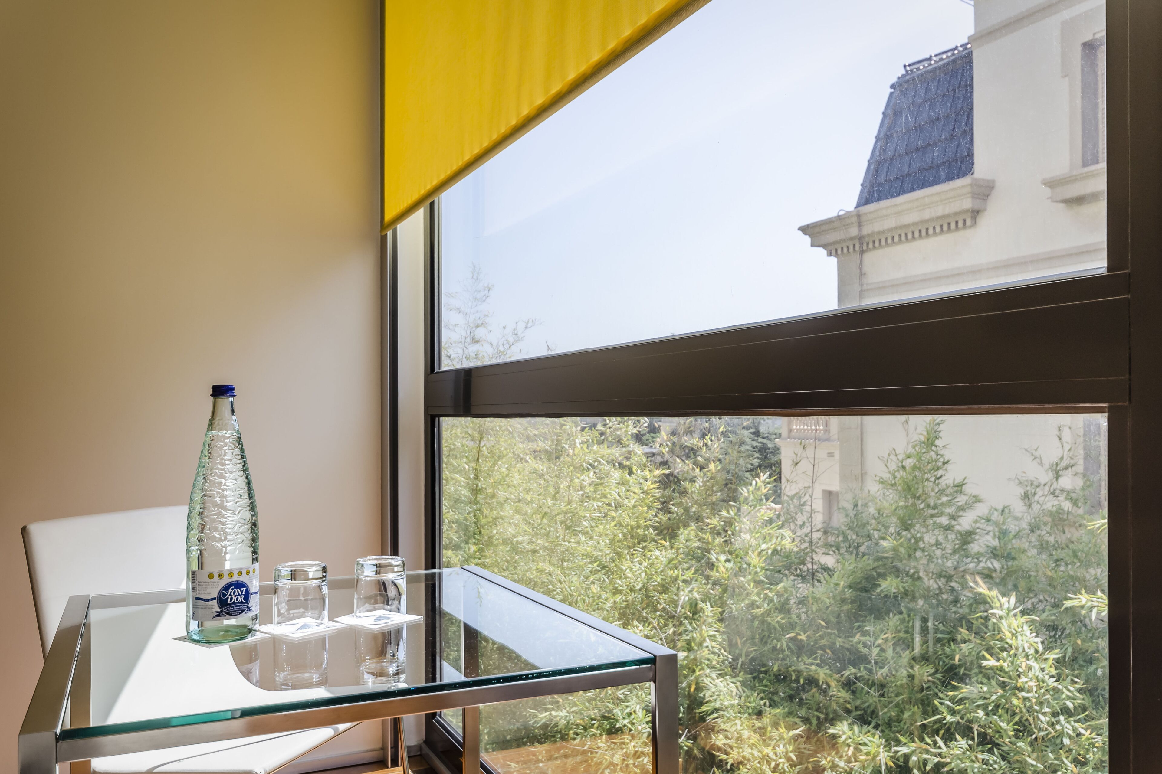 Een glazen tafel met een fles en glazen, een geel gordijn en een uitzicht op een gebouw en groen.