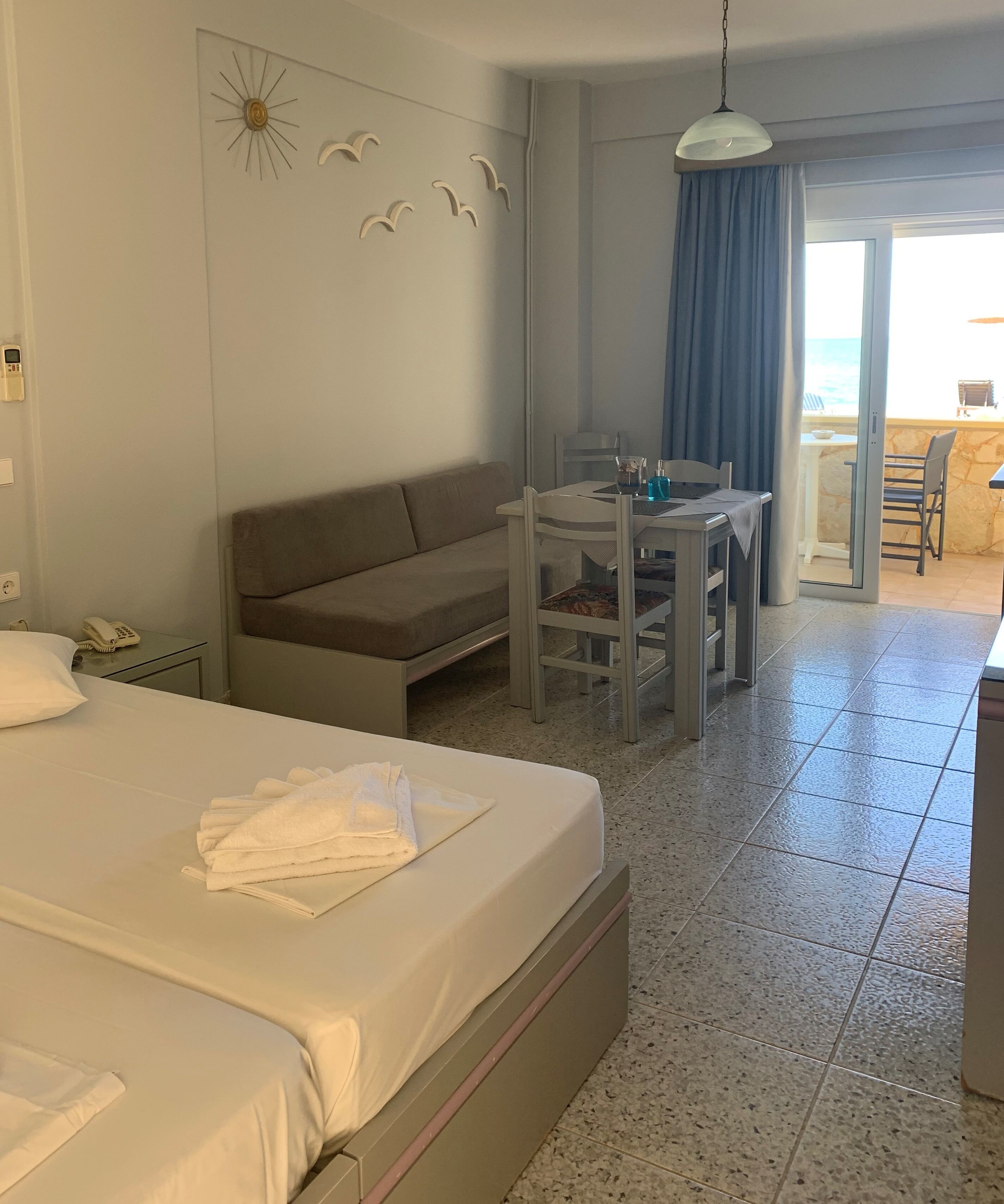 Ein Hotelzimmer mit Bett, Sofa, Esstisch mit Stühlen, einem kleinen Tisch mit Lampe und Blick ins Freie.