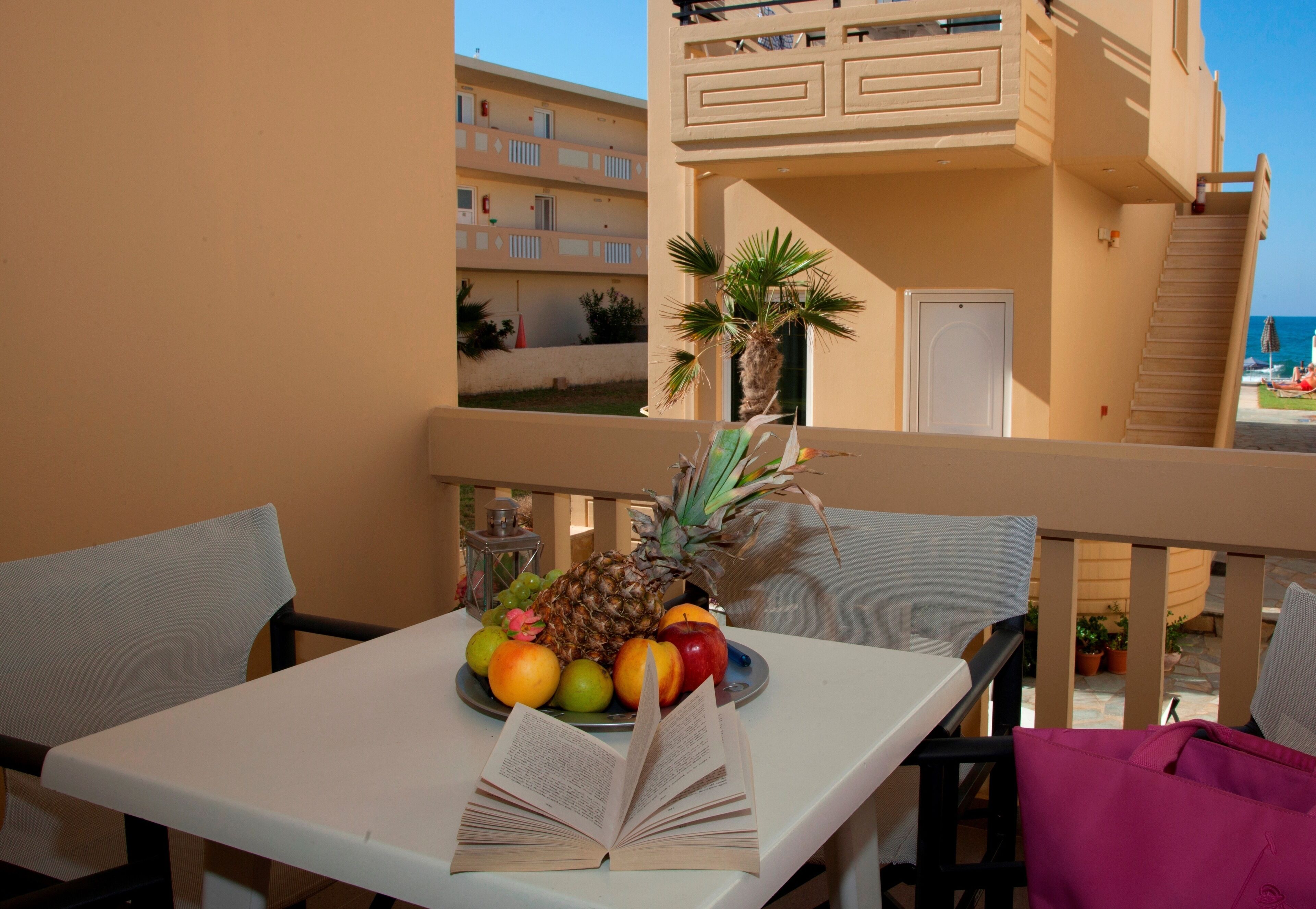 A balcony with a table set with fruits and an open book, overlooking a building with balconies and a clear sky.