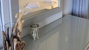 Habitación de hotel con un espejo grande que refleja una cama bien hecha, un jarrón de vidrio con pampa seca y un adorno de madera con forma de corazón sobre una superficie reflectante.