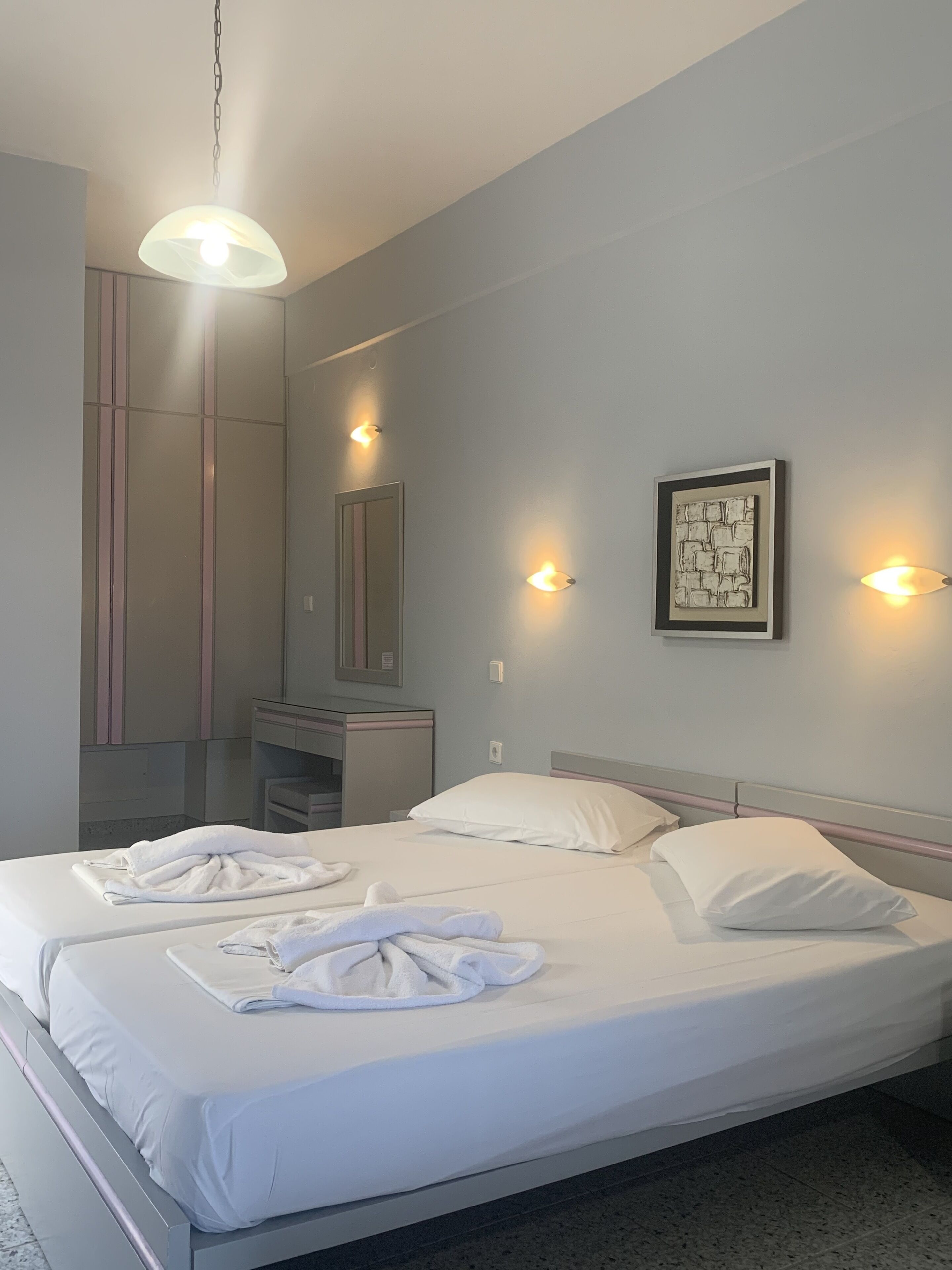 A modern hotel room with a large bed, bedside tables, a vanity with a mirror, and wall-mounted lighting.