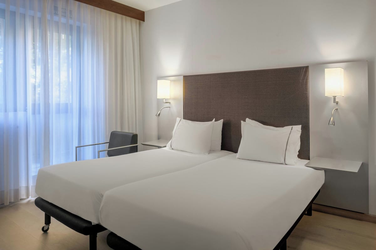 A hotel room with a large bed, two bedside tables, a chair, and a window with sheer curtains.