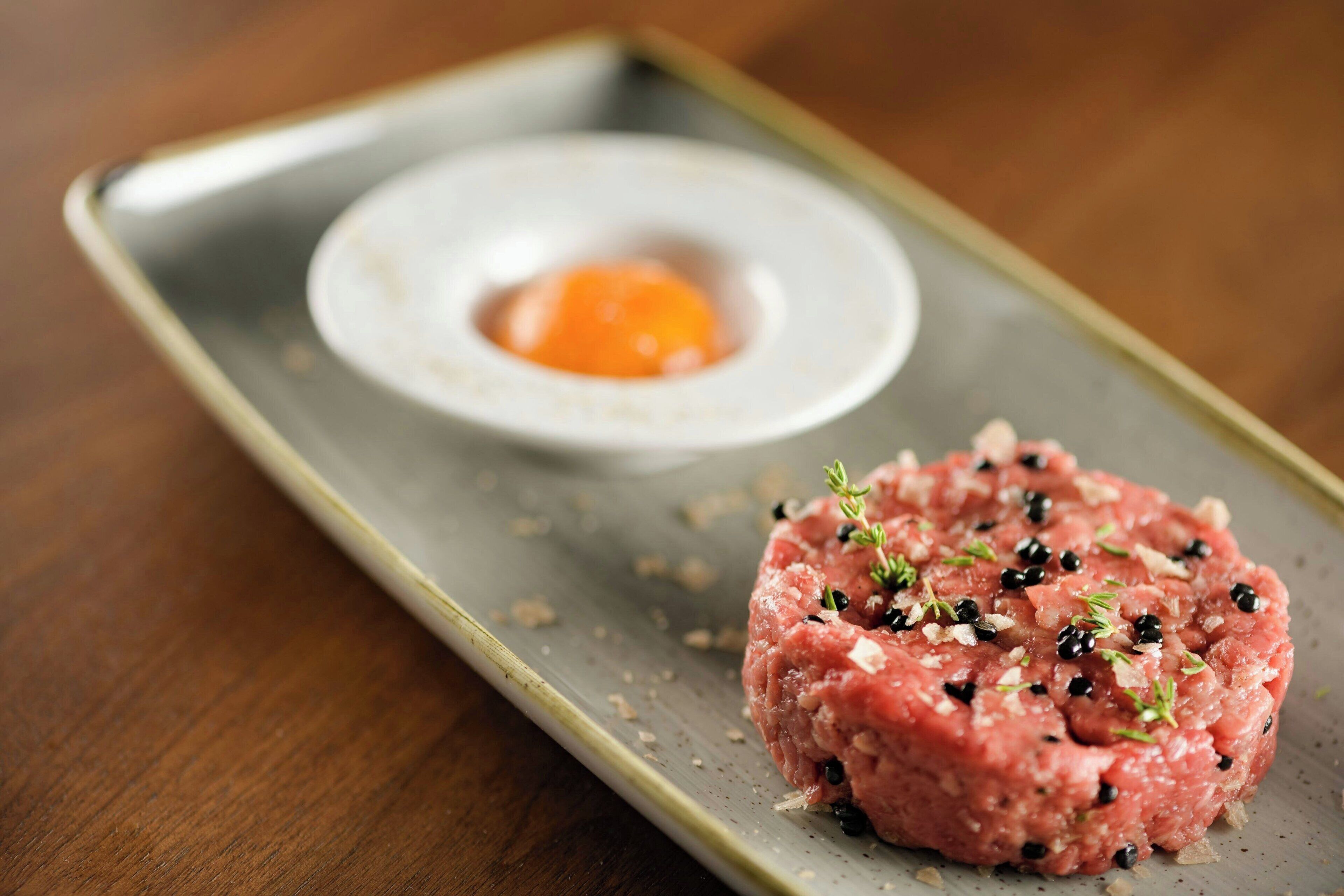 Un plato de carne molida cruda adornado con hierbas y pimienta negra, servido en un plato rectangular con un recipiente pequeño de yema de huevo.