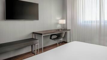 A modern hotel room with a large bed, a desk with a chair, a TV mounted on the wall, and a window with sheer curtains.