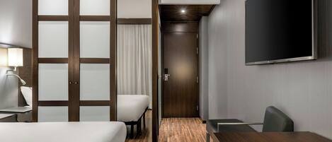 Habitación de hotel con cama, escritorio, silla, televisor y puerta corrediza con paneles de vidrio satinado.