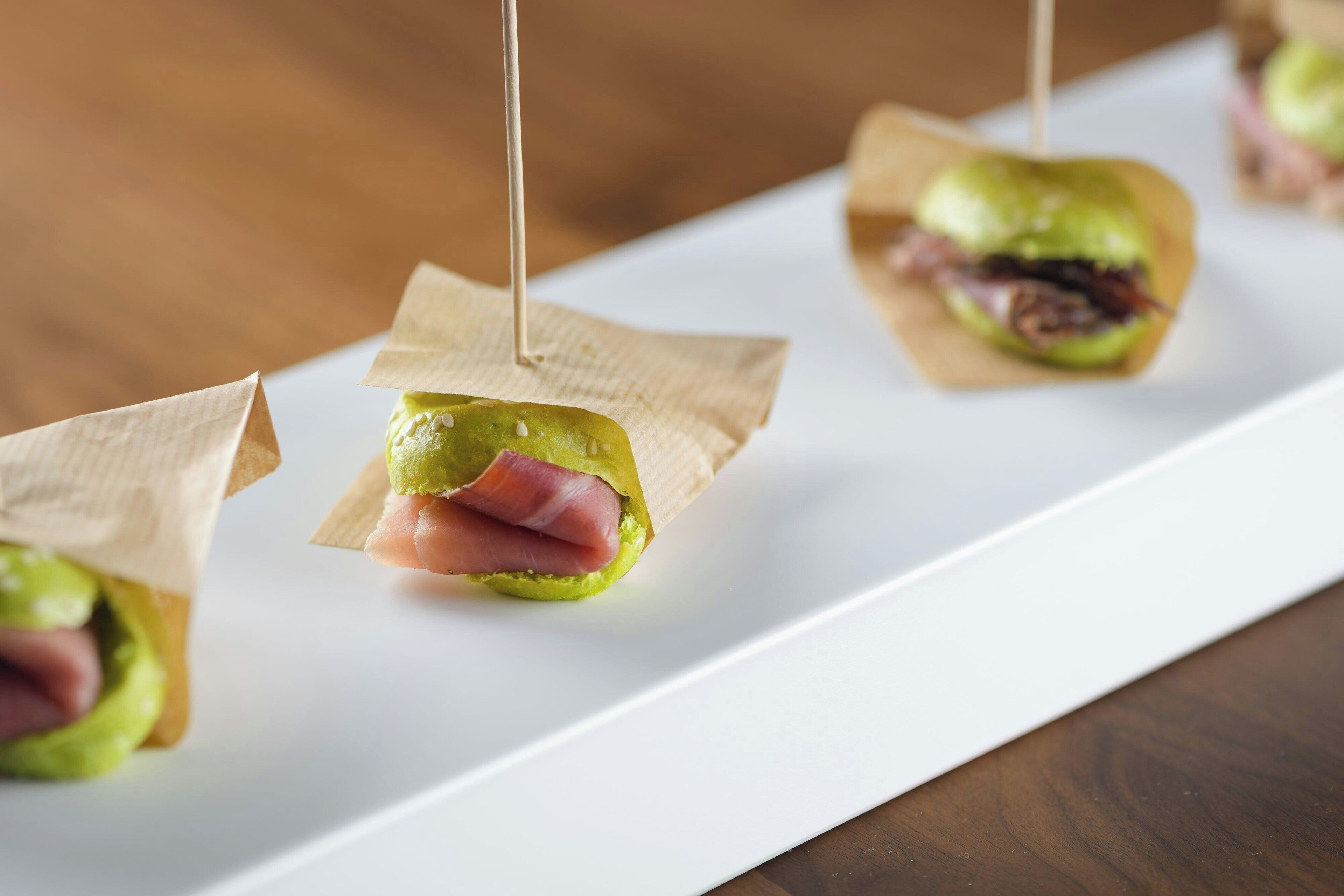 A white rectangular plate with three food items, each wrapped in brown paper and held by toothpicks.