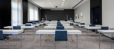 A conference room with multiple tables and chairs, large windows with curtains, and a projector screen at the front.