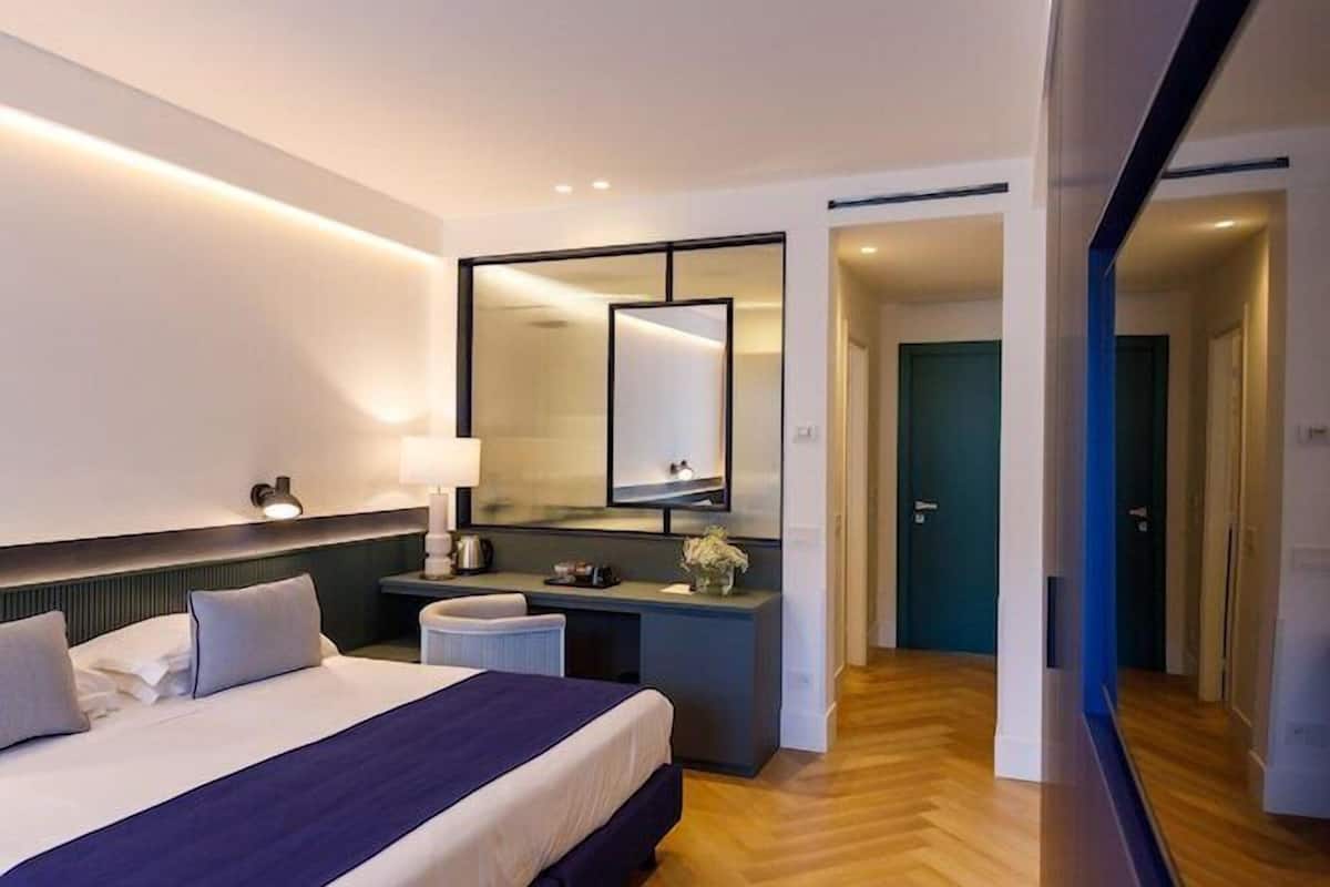 Ein modernes Hotelzimmer mit einem großen Bett, Nachttisch, Spiegel und Blick auf den Flur.