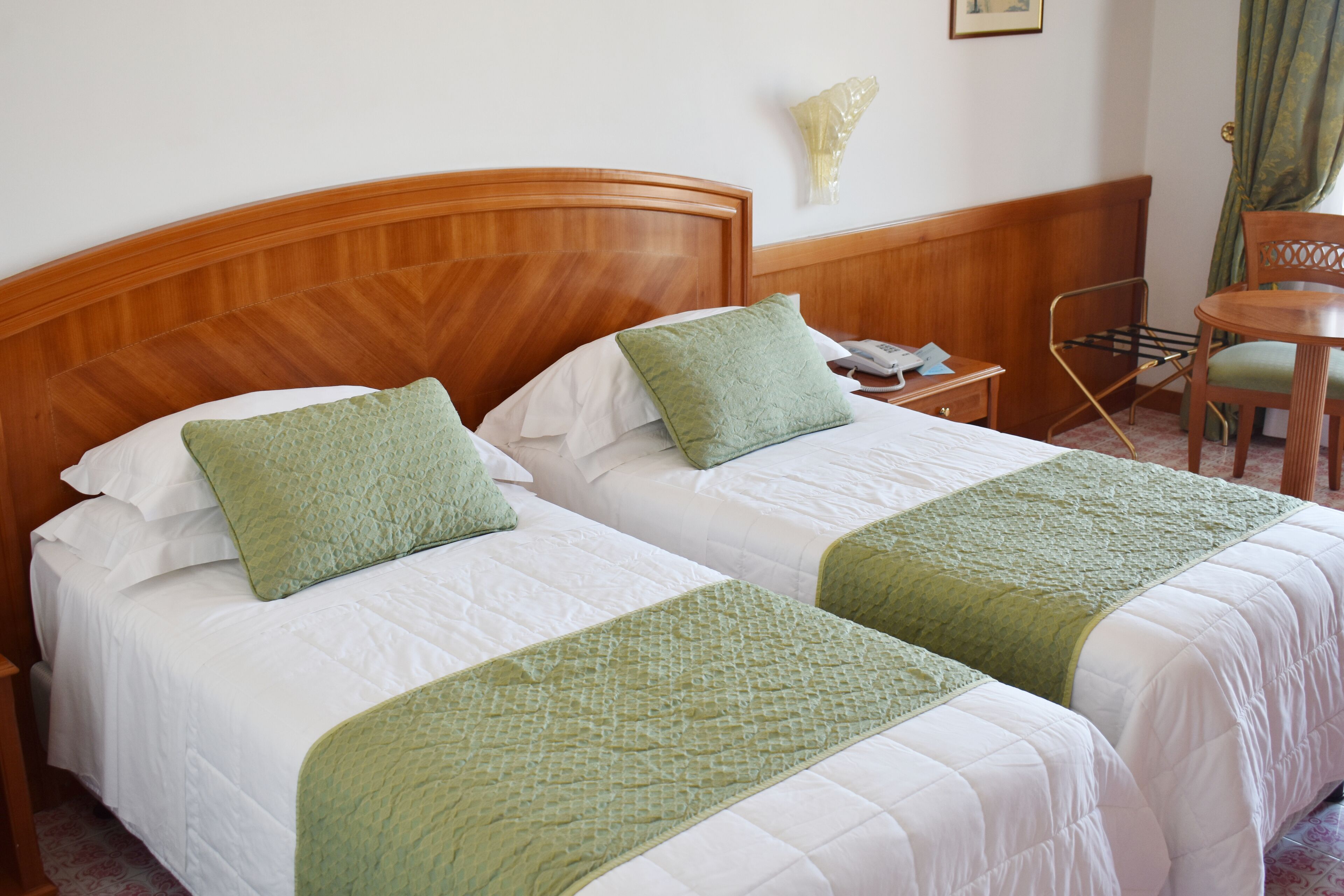 Habitación de hotel con dos camas, cada una con almohadas y colcha verdes. Tiene un cabecero de madera, una mesita con una silla y una lámpara de pared.