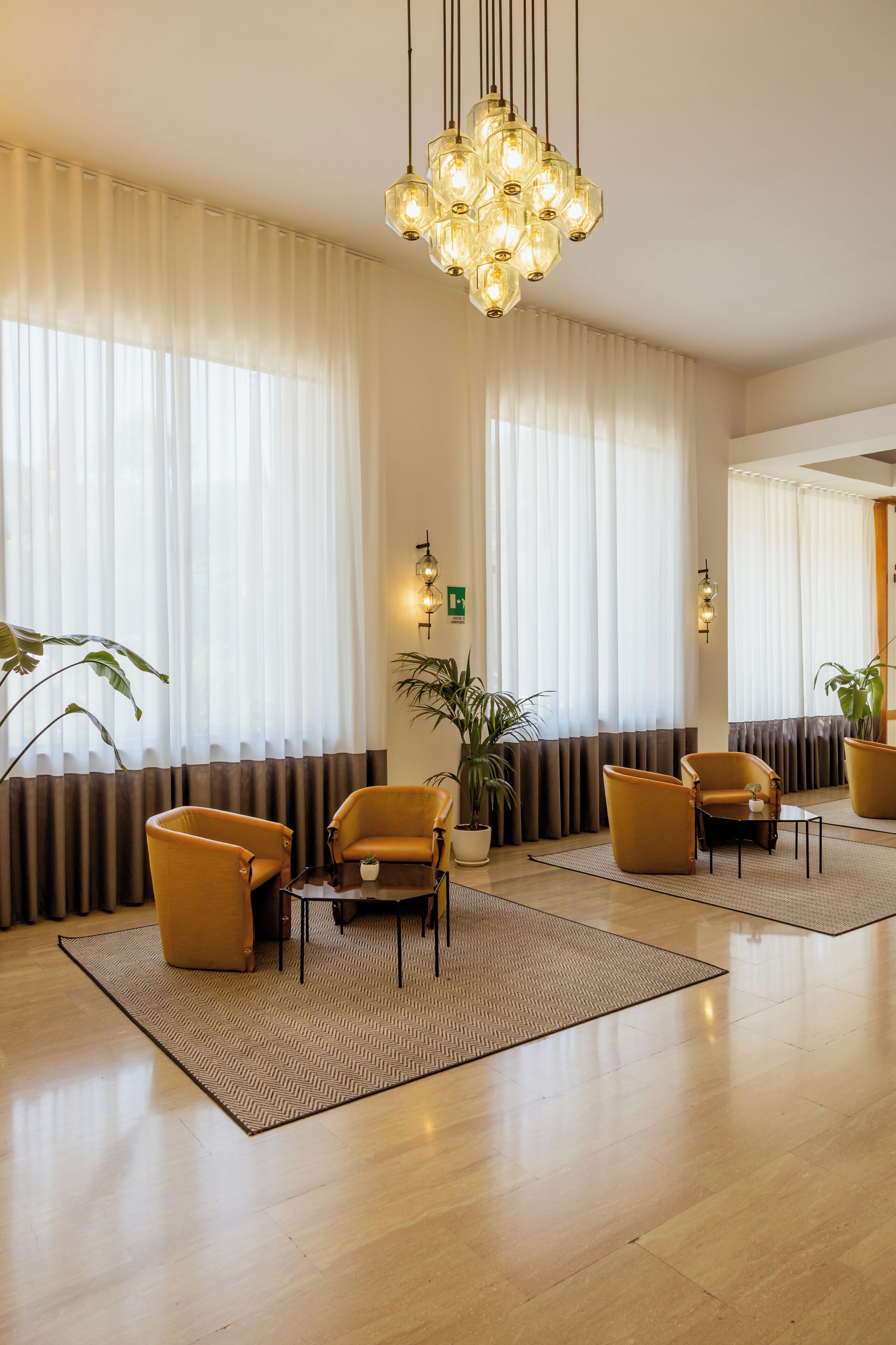 Een hotellobby met een kroonluchter, grote ramen met gordijnen en verschillende fauteuils.