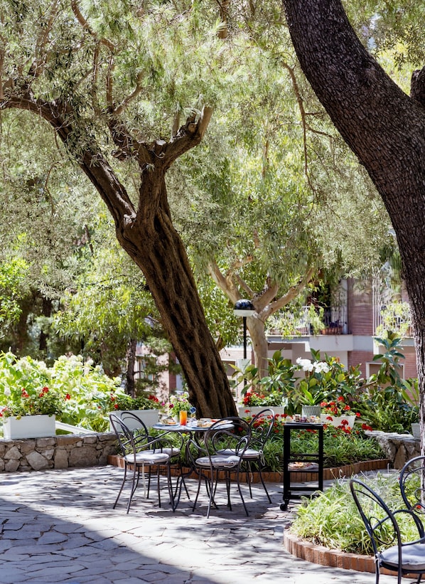 Un patio exterior con suelo de piedra, sillas y mesas de metal, rodeado de abundante vegetación y plantas con flores.