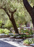 Un patio esterno con pavimento in pietra, sedie e tavoli in metallo, circondato da vegetazione lussureggiante e piante fiorite.