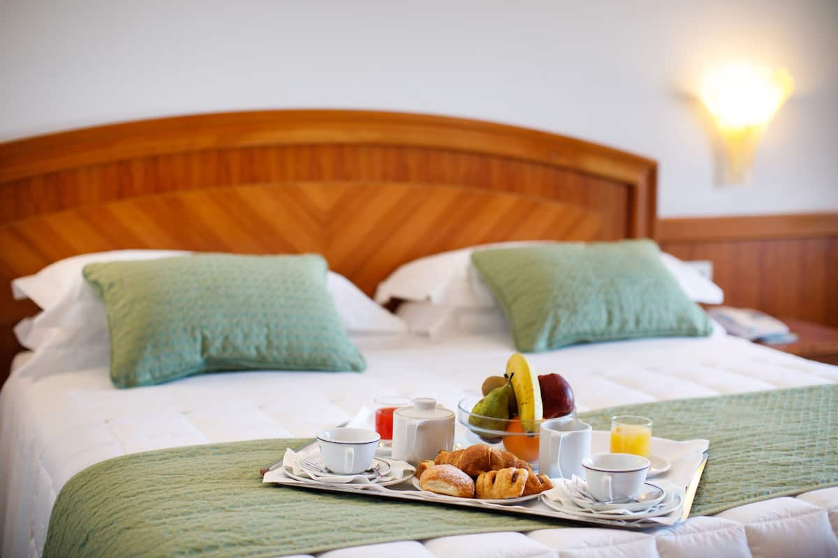 Ein ordentlich bezogenes Bett mit einer grünen Tagesdecke, ein Tablett mit Frühstücksartikeln, darunter Croissants, Obst und Getränke, und ein hölzernes Kopfteil.