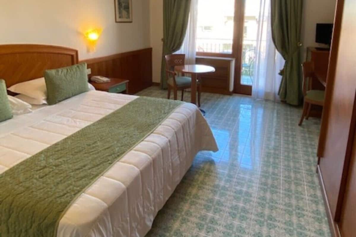 Une chambre d’hôtel comprenant un lit, un bureau, une chaise, une télévision et un balcon avec des rideaux.