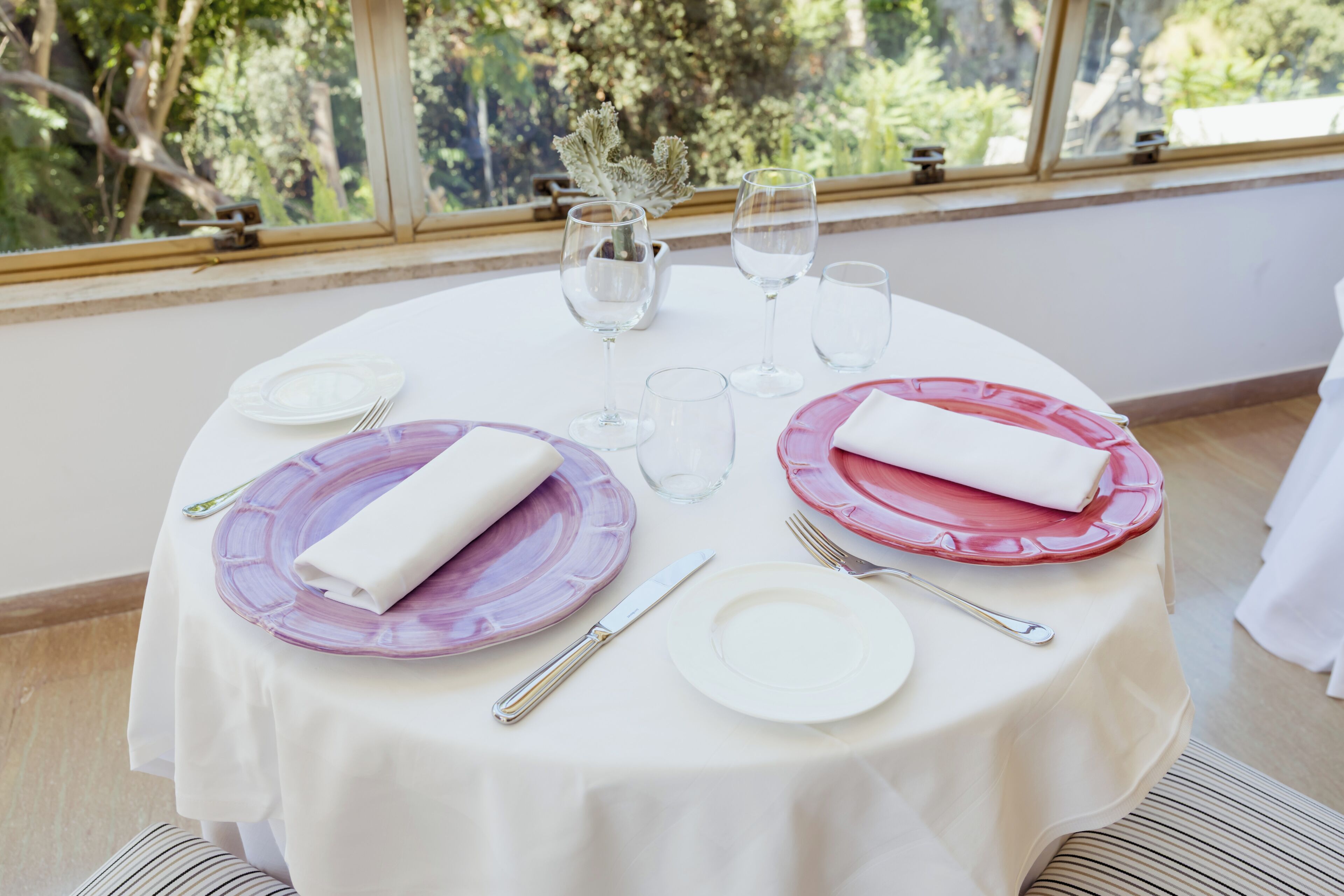 A round table set for two with white and purple plates, silverware, and clear glasses.
