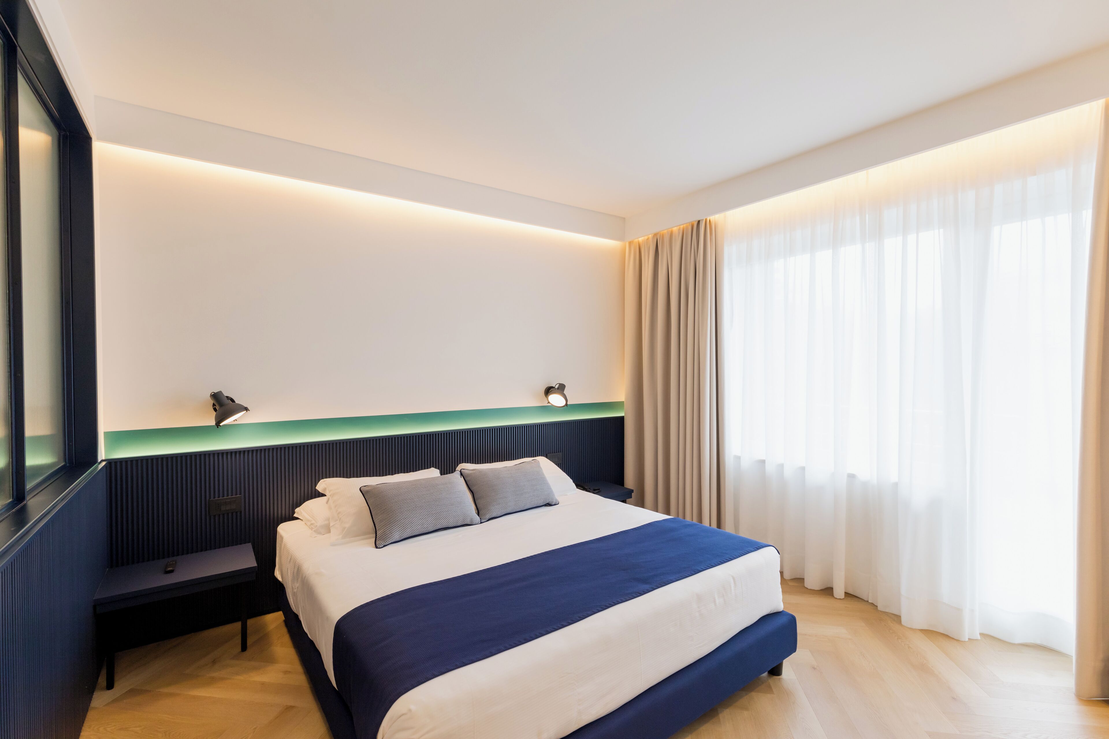 A modern hotel room with a large bed, bedside tables, a wall-mounted lamp, and a large window with curtains.