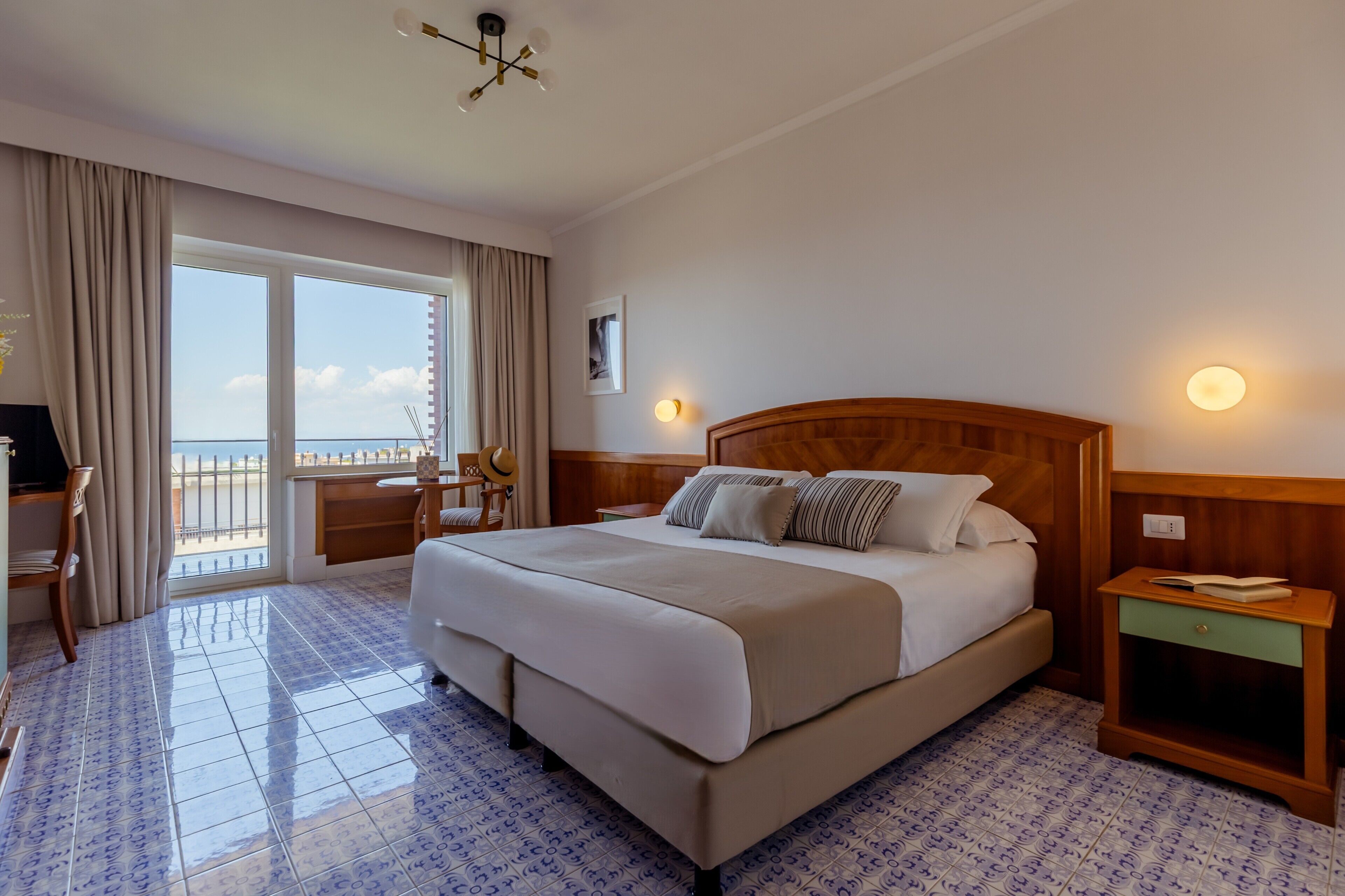 Camera d'albergo con un letto grande, una scrivania, una sedia, un comodino e vista mare dalla finestra.