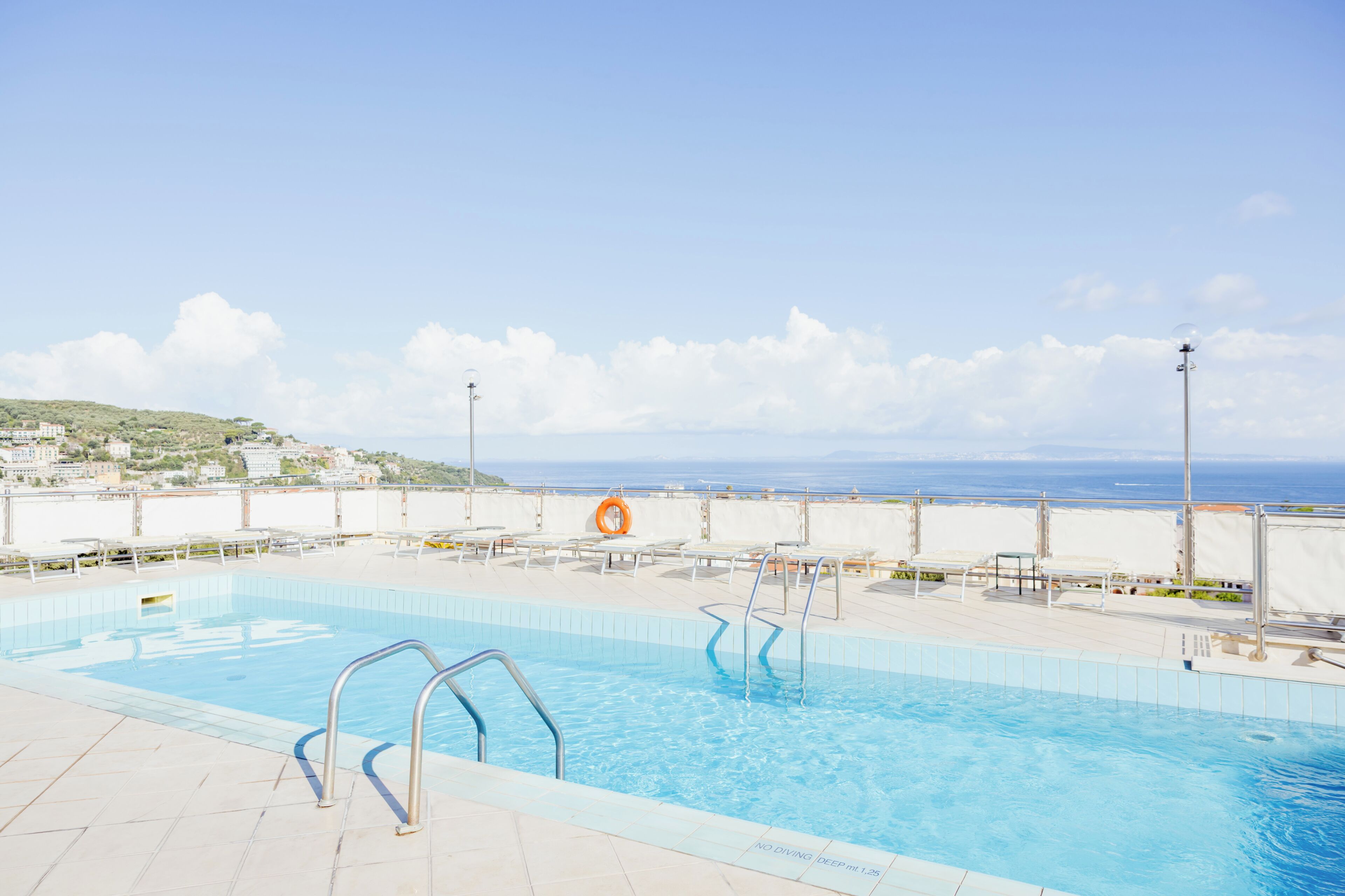 A swimming pool with clear blue water, a lifebuoy, and a view of the sea and buildings in the background.