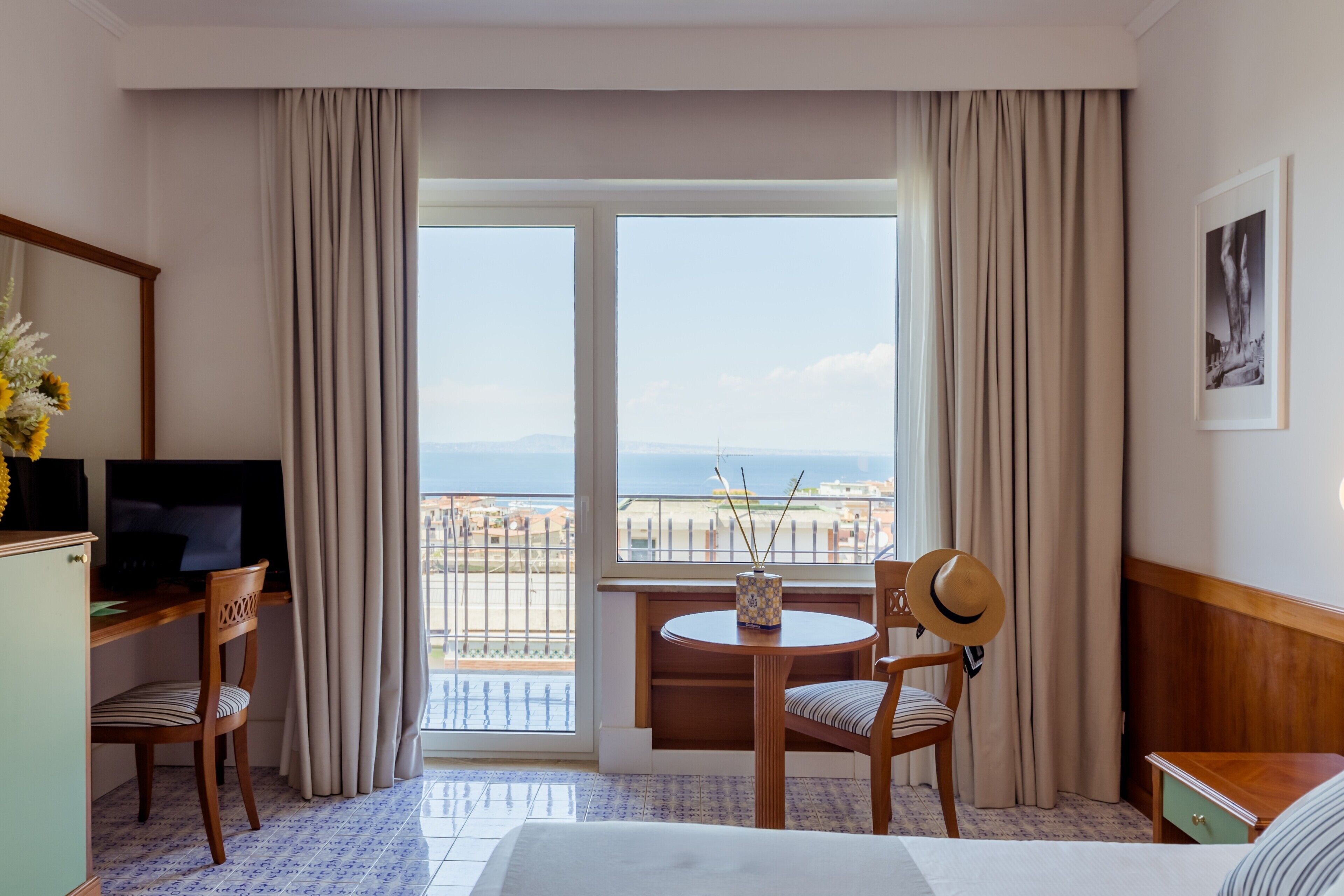 Habitación de hotel con cama, escritorio con televisor, silla, mesita con un jarrón y balcón con vista al mar.