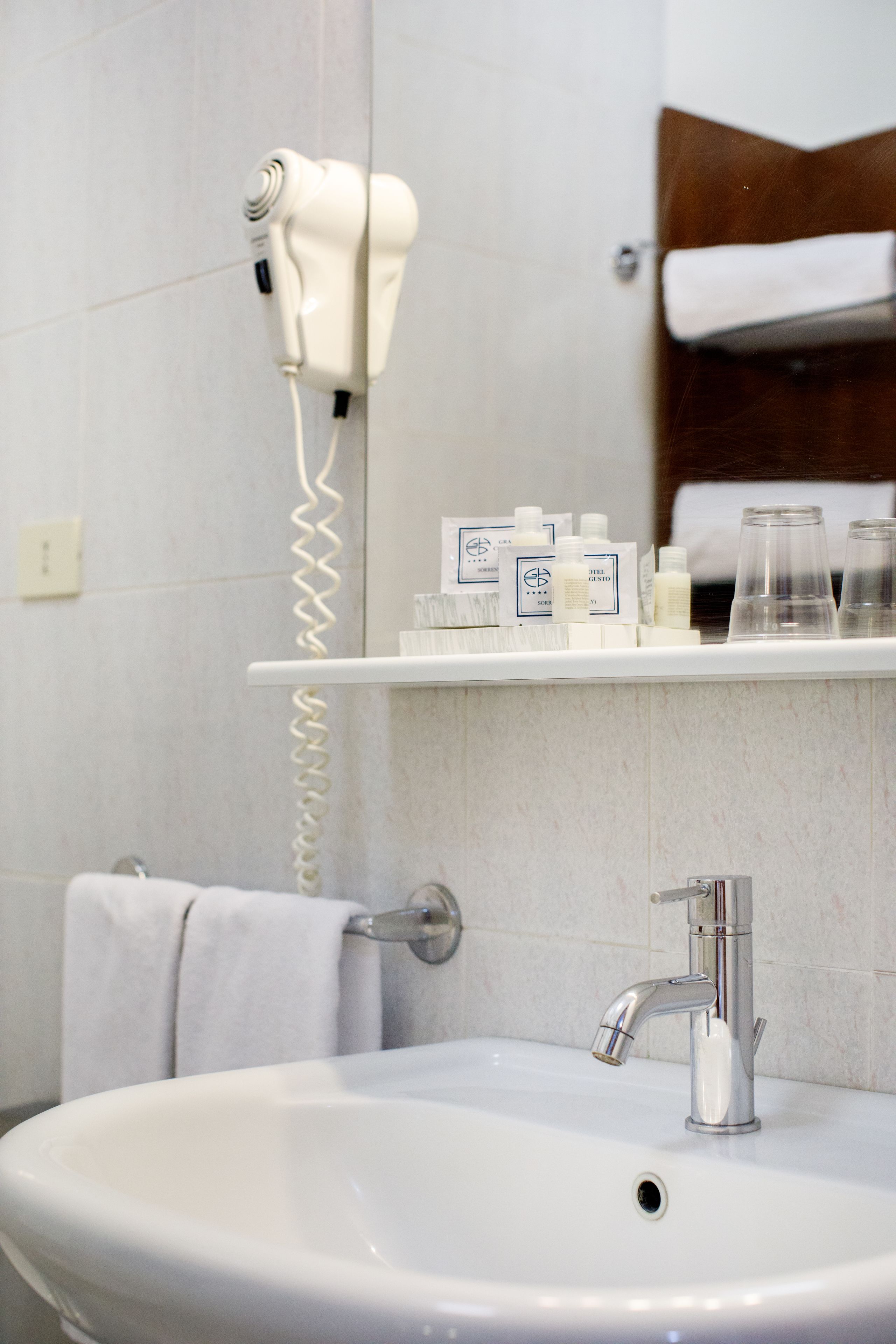 Une salle de bain avec un lavabo blanc, un sèche-cheveux posé sur un support mural et une étagère contenant des articles de toilette.