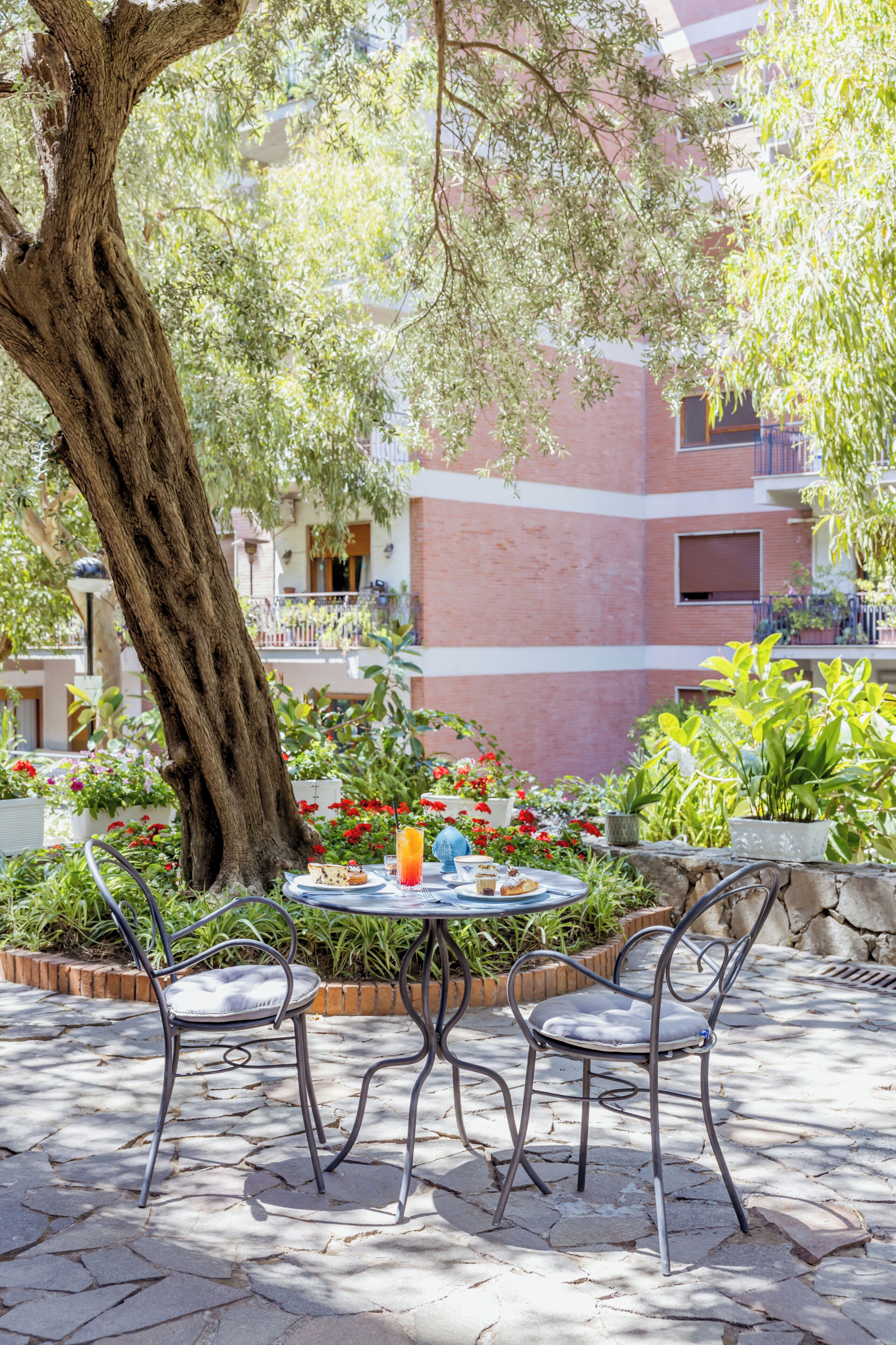 An outdoor seating area with a round table and metal chairs, surrounded by potted plants and a large tree.