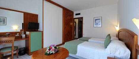 A hotel room with a bed, a desk with a television, a chair, a table with a flower arrangement, and a bedside table with a phone.
