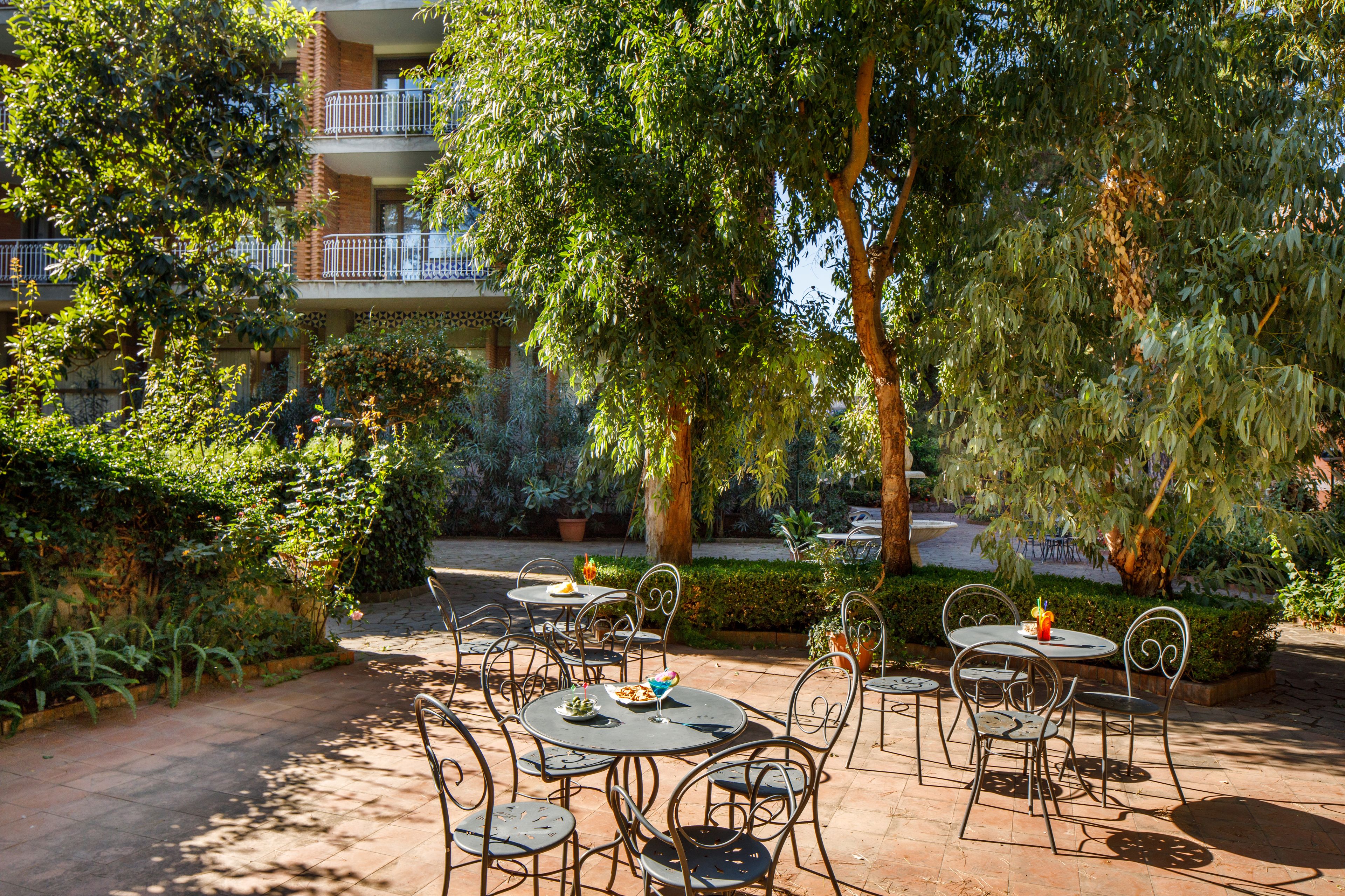 Patio esterno con tavoli rotondi e sedie in metallo, circondato da rigogliosa vegetazione e alberi.