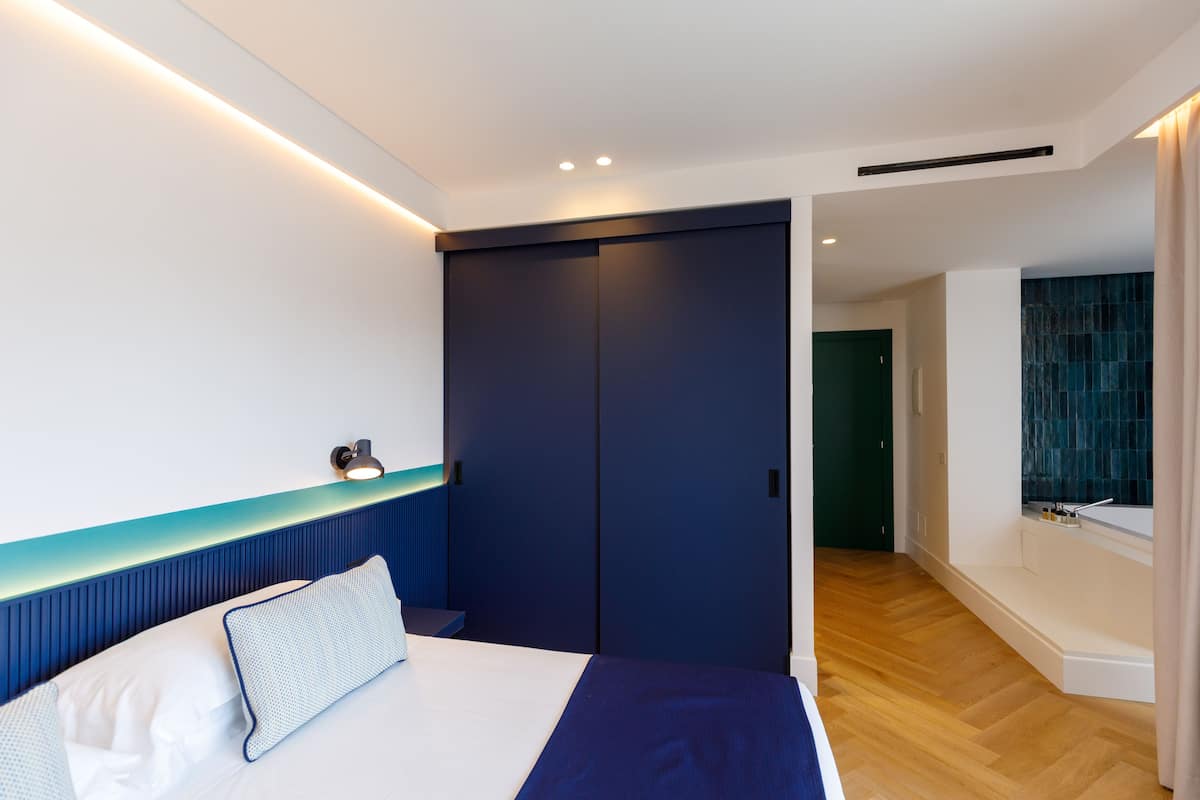 Une chambre moderne avec un lit, une table de chevet, une armoire bleu foncé et une salle de bain dont un mur est recouvert de carreaux bleus.