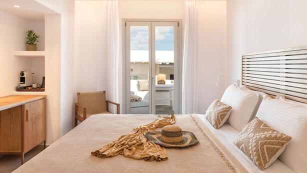 A bedroom with a bed, a wooden headboard, a bedside table, a chair, a window with curtains, and a view of the outside. - Elena Hotel (Mykonos)