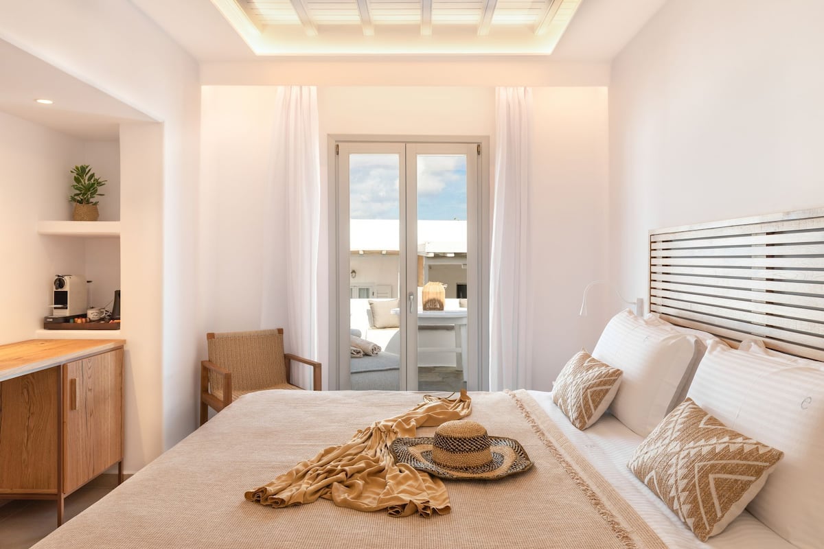 A bedroom with a bed, a wooden headboard, a bedside table, a chair, a window with curtains, and a view of the outside.