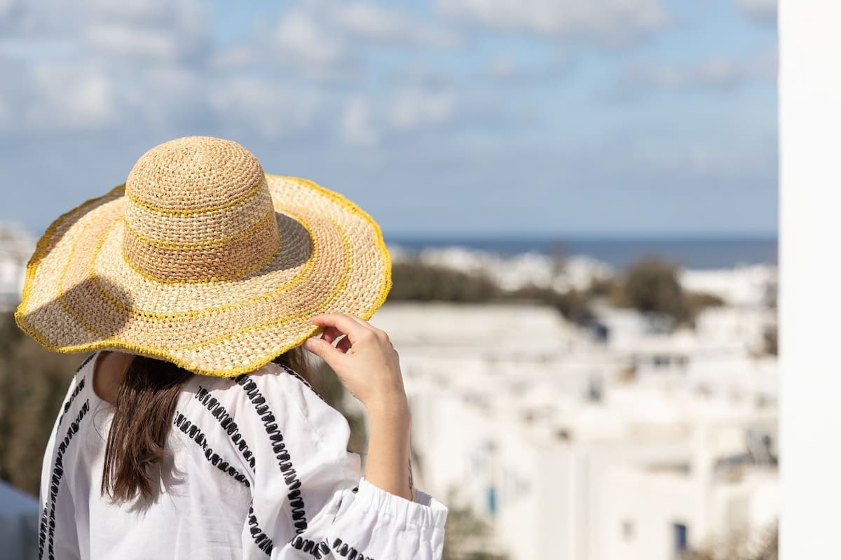 A person wearing a straw hat and a white shirt with black patterns, standing with a view of white buildings and a blue sky.