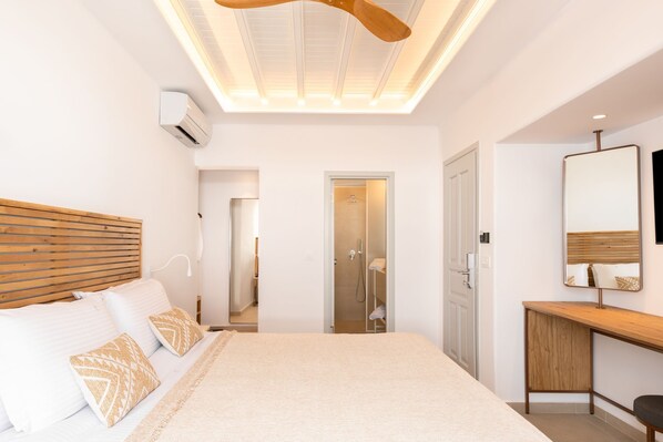 A modern bedroom with a wooden headboard, a large bed with patterned pillows, a bedside table, a mirror, and a walk-in closet. - Elena Hotel (Mykonos)