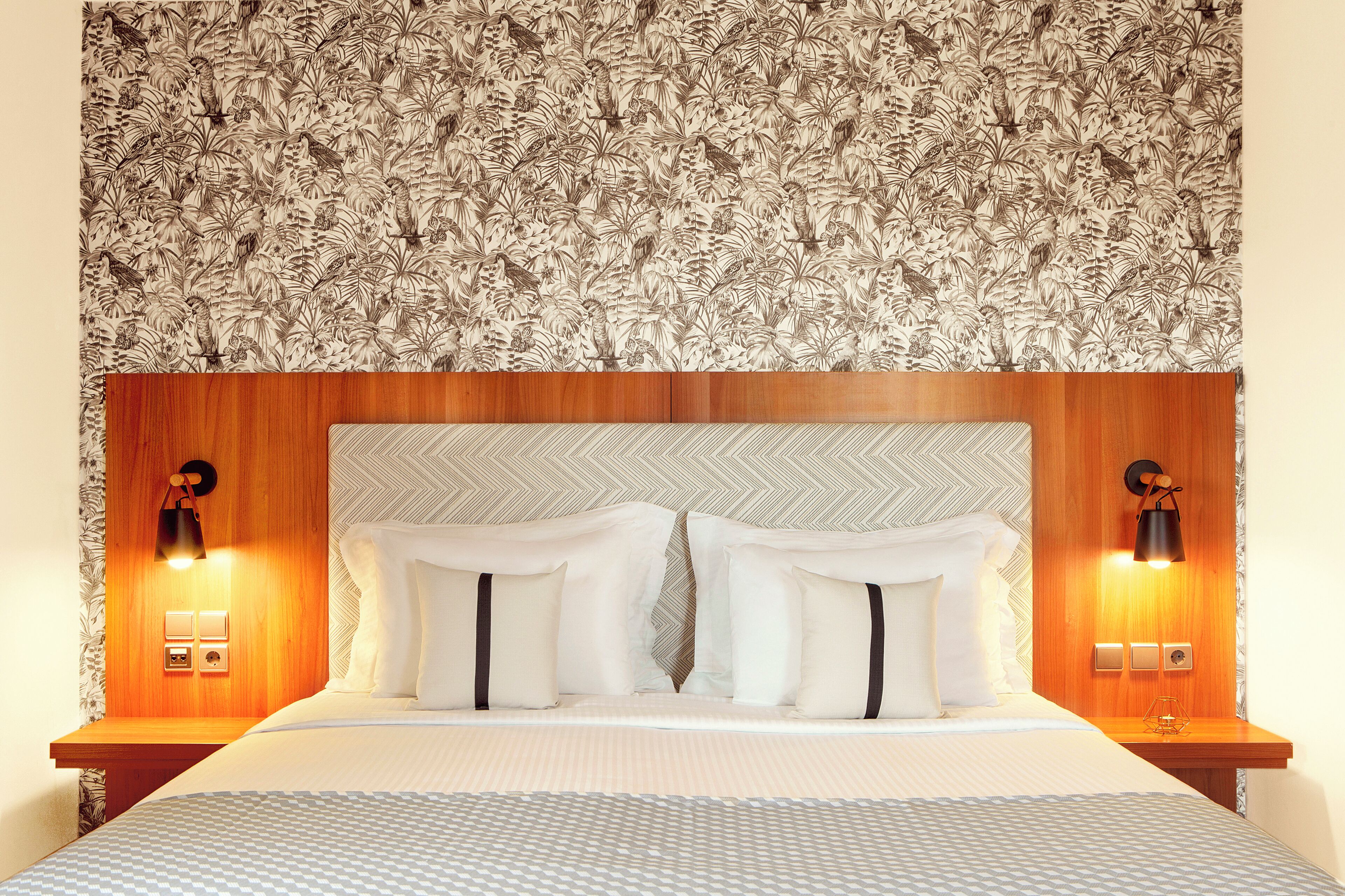 Habitación de hotel con una cama grande, dos mesitas de noche con lámparas, un cabecero de madera y un estampado floral en el papel tapiz.