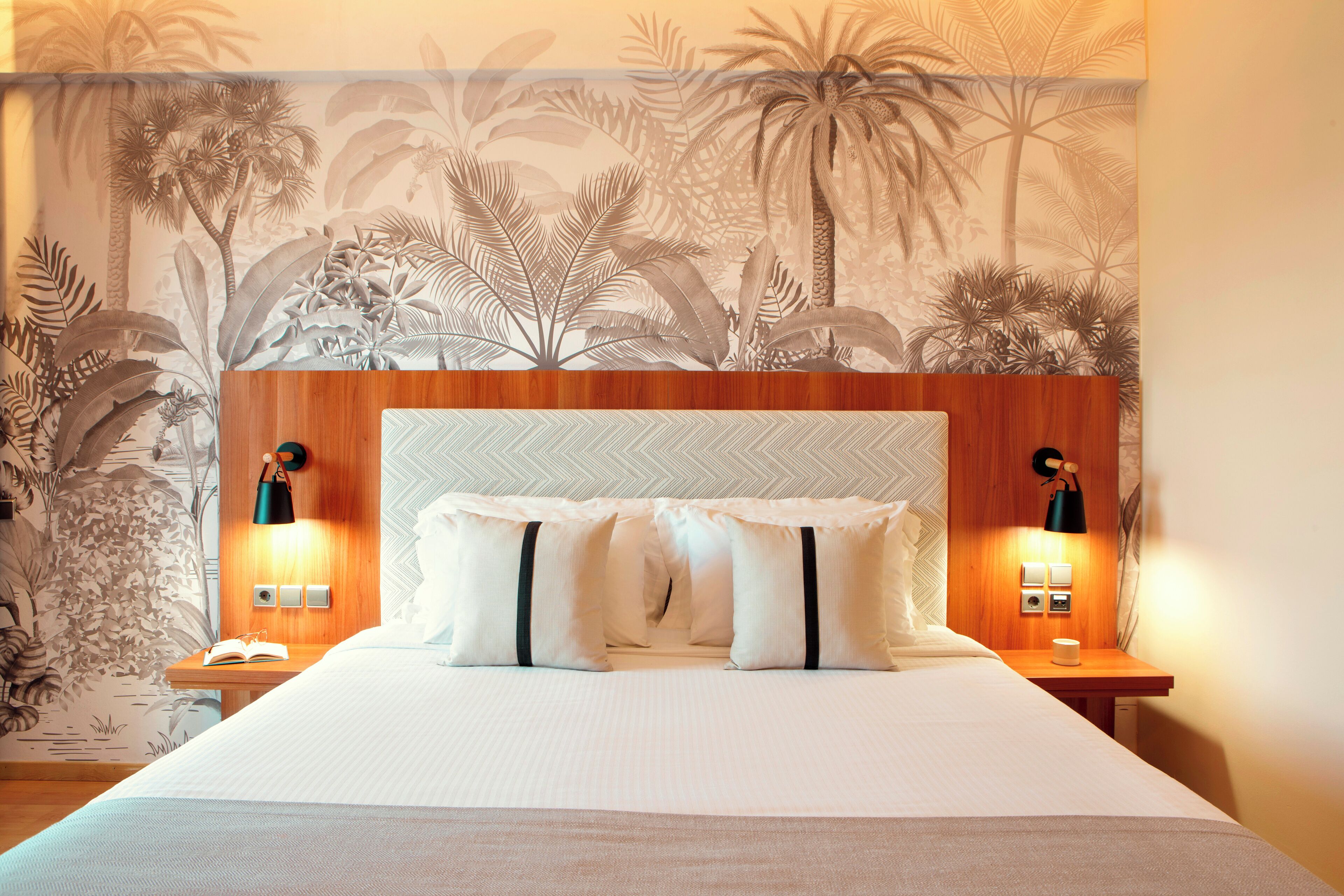 Habitació d'hotel amb un llit gran, dues taules de nit, un taulell, una paret amb un mural tropical i dues làmpades penjades a la pared.